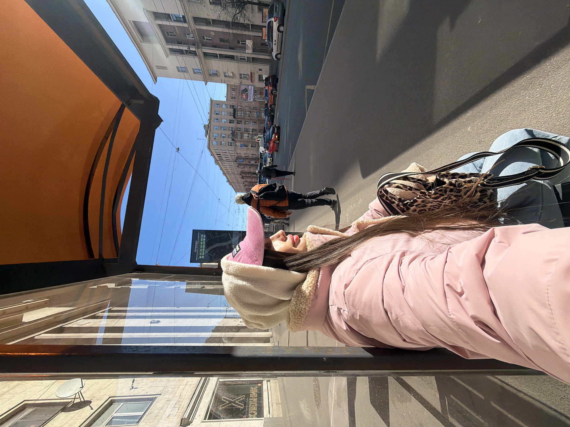 The image depicts a young woman at a bus stop in Saint Petersburg, Russia. She is wearing a pink hooded coat and a pink baseball cap, standing beside a pane of glass at a bus stop. Reflected in the glass are buildings, a satellite dish, and a sign in Russian that includes the word "ХЛЕБНИК". Outside the bus stop, pedestrians walk along the sidewalk, and cars are parked on the street. A large billboard is visible in the background, between the buildings. The sky is bright blue.
