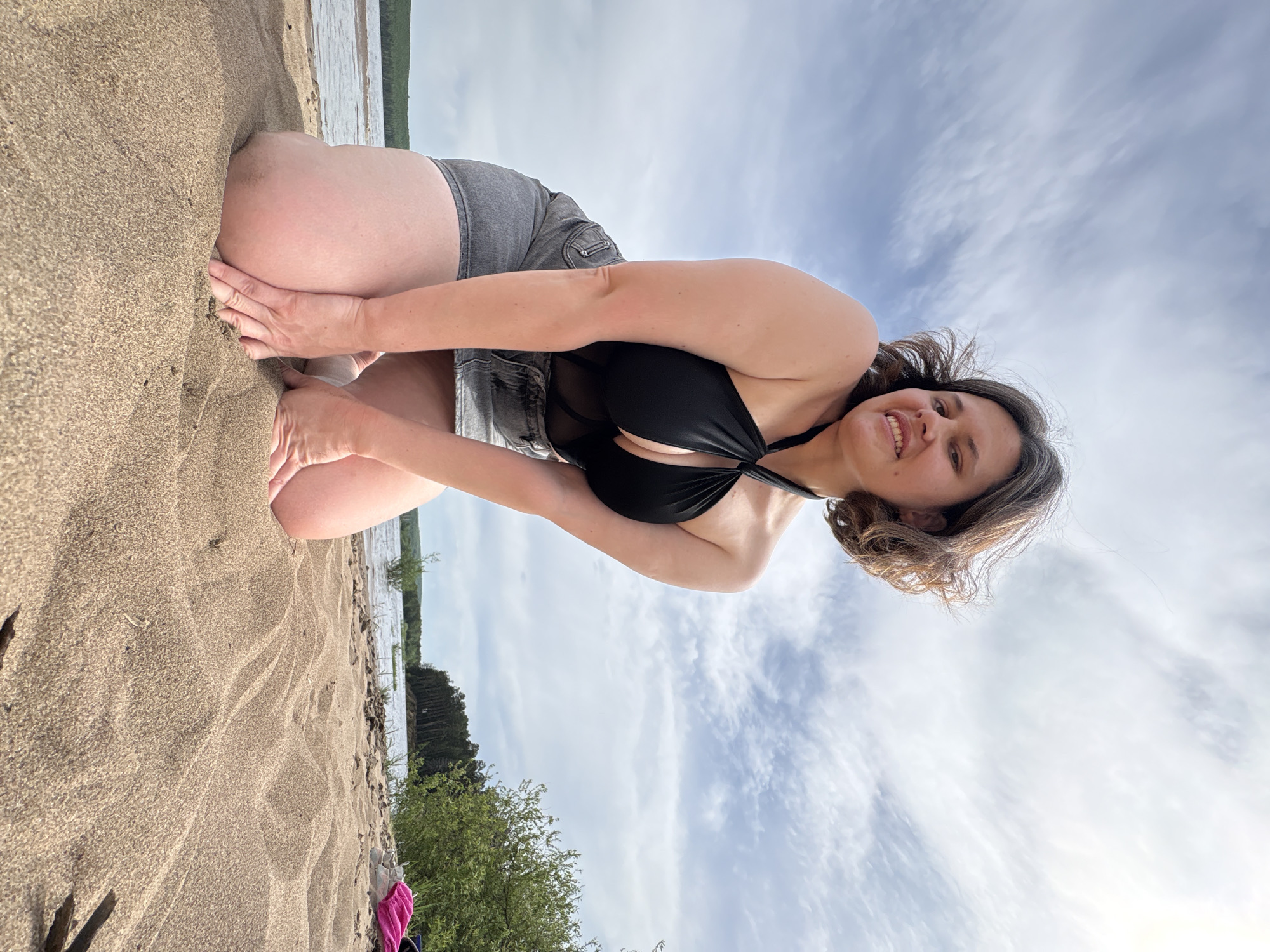 The image shows a woman kneeling on a sandy beach, possibly in the city of Chaikovsky, Russia, given the context. She is wearing a black swimsuit top and grey denim shorts. The woman is leaning forward, with her hands touching the sand. She is smiling and looking at the camera. In the background is a body of water, possibly a river or lake, and a cloudy sky. To the left of the frame are some green bushes.
