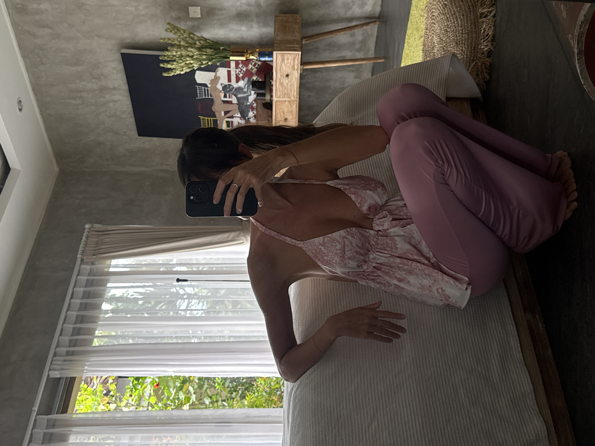The image shows a woman taking a mirror selfie indoors. She is seated on a bed or daybed, positioned with her back partially to the viewer, but her head is turned to the left as she holds up a dark-cased smartphone to capture her reflection, obscuring her face. Her long, dark hair cascades down her back. She wears a light-colored top with a pink or white pattern and thin straps, paired with form-fitting, light purple/mauve pants. Her legs are bent, and her bare feet are visible towards the right side of the frame. A thin bracelet is on her right wrist, and a ring is on her left hand's ring finger.
The setting appears to be a modern room, consistent with a location like North Kuta, Indonesia. A large window on the left side of the frame features horizontal blinds and offers a view of lush green foliage and bright daylight, indicating it is daytime and likely sunny outside. The room's walls have a textured, gray concrete finish. In the background, a dark rectangular piece of wall art depicting abstract figures hangs above a rustic wooden console table, which holds a tall arrangement of light-colored, spiky flowers or dried plants. The bed she is sitting on has a ribbed white or light-colored mattress cover. The floor to the right appears dark. There is no visible text in the image.
