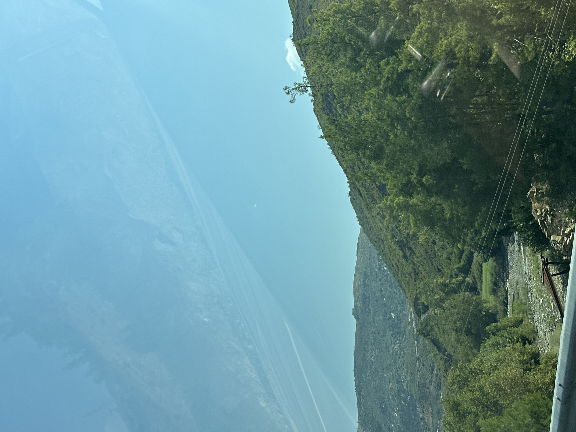 The image depicts a natural landscape dominated by lush green hills and a clear blue sky. A rocky riverbed is visible in the lower part of the image, with a few sparse trees lining its banks. Power lines stretch across the scene, strung between unseen poles. The terrain suggests a rural or mountainous area, possibly within the vicinity of Manavgat, Turkey, given the characteristic Mediterranean vegetation and topography. There are no people or specific structures that would indicate a defined urban setting within Manavgat itself. The scene is serene and natural, highlighting the environmental features of the region.