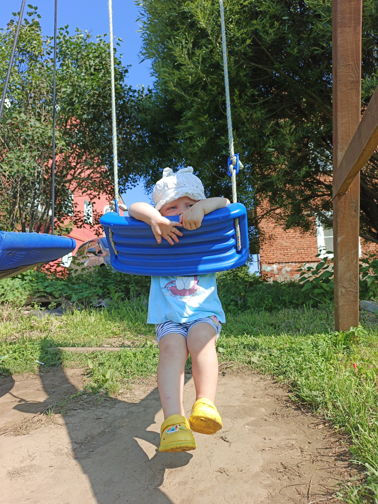 A child is sitting on a blue swing in a backyard in Medvedyovka, Russia. The child is wearing a white hat, a light blue t-shirt with a cartoon character, and striped shorts. Yellow shoes are on the child's feet. The swing is suspended from a wooden frame. In the background, there is a red brick building, green trees, and green grass. The sky is blue. The overall impression is that of a sunny day in a rural or suburban setting.