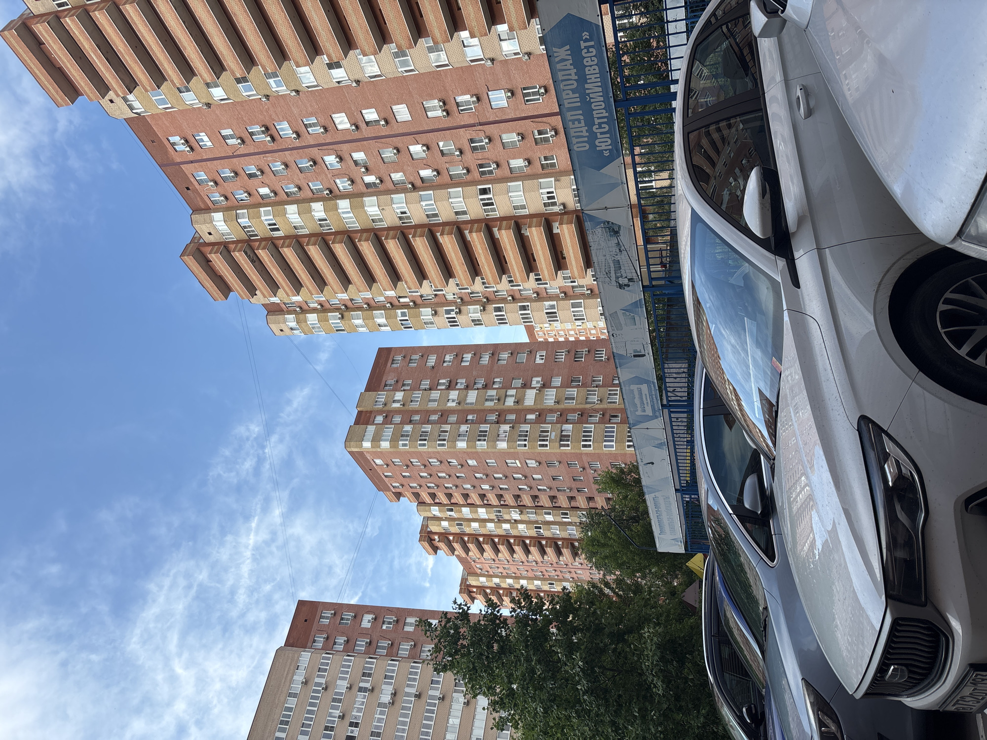 The image captures a street scene in Krasnodar, Russia, dominated by tall, multi-story residential buildings. The buildings are constructed with a mix of light and reddish-brown brick, featuring numerous windows and balconies, suggesting a densely populated urban environment. In the foreground, several cars are parked, with a white car prominently displayed on the right side of the frame.
The sky is blue with scattered white, wispy clouds, indicating fair weather, likely during the daytime. A banner with Russian text is visible on a blue fence behind the buildings, reading "ОТДЕЛ ПРОДАЖ «ЮгСтройИнвест»" which translates to "Sales Department 'YugStroyInvest'". This suggests the buildings might be part of a new development or construction project.
The perspective is from a low angle, looking upwards, emphasizing the height of the buildings and the expanse of the sky. Green trees are interspersed between the buildings, adding a touch of nature to the urban landscape. The overall scene depicts a typical day in a residential area, with cars parked and buildings standing tall under a clear sky. No people are clearly visible in the image.