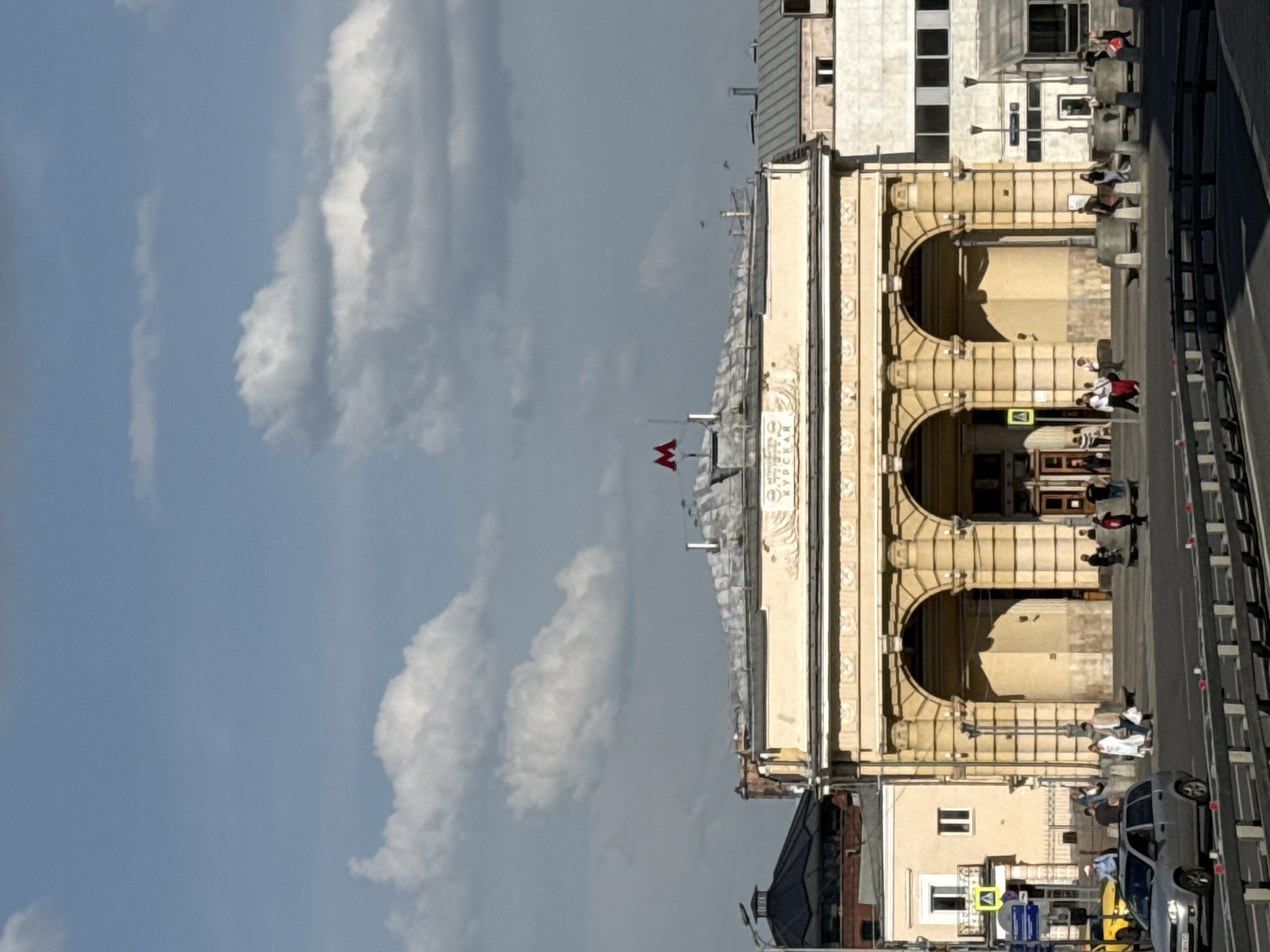 The image depicts an architectural scene in Moscow, Russia, featuring the Kurskaya metro station building. The building has a classical design with arched entrances and is constructed primarily of beige or light yellow stone. The background includes a mix of partly cloudy sky and other buildings, some of which are taller and more modern. People are seen on the sidewalk and crossing the road in front of the station, indicating pedestrian traffic. Cars and possibly other vehicles are visible on the road. The image suggests a daytime scene in a busy urban area.