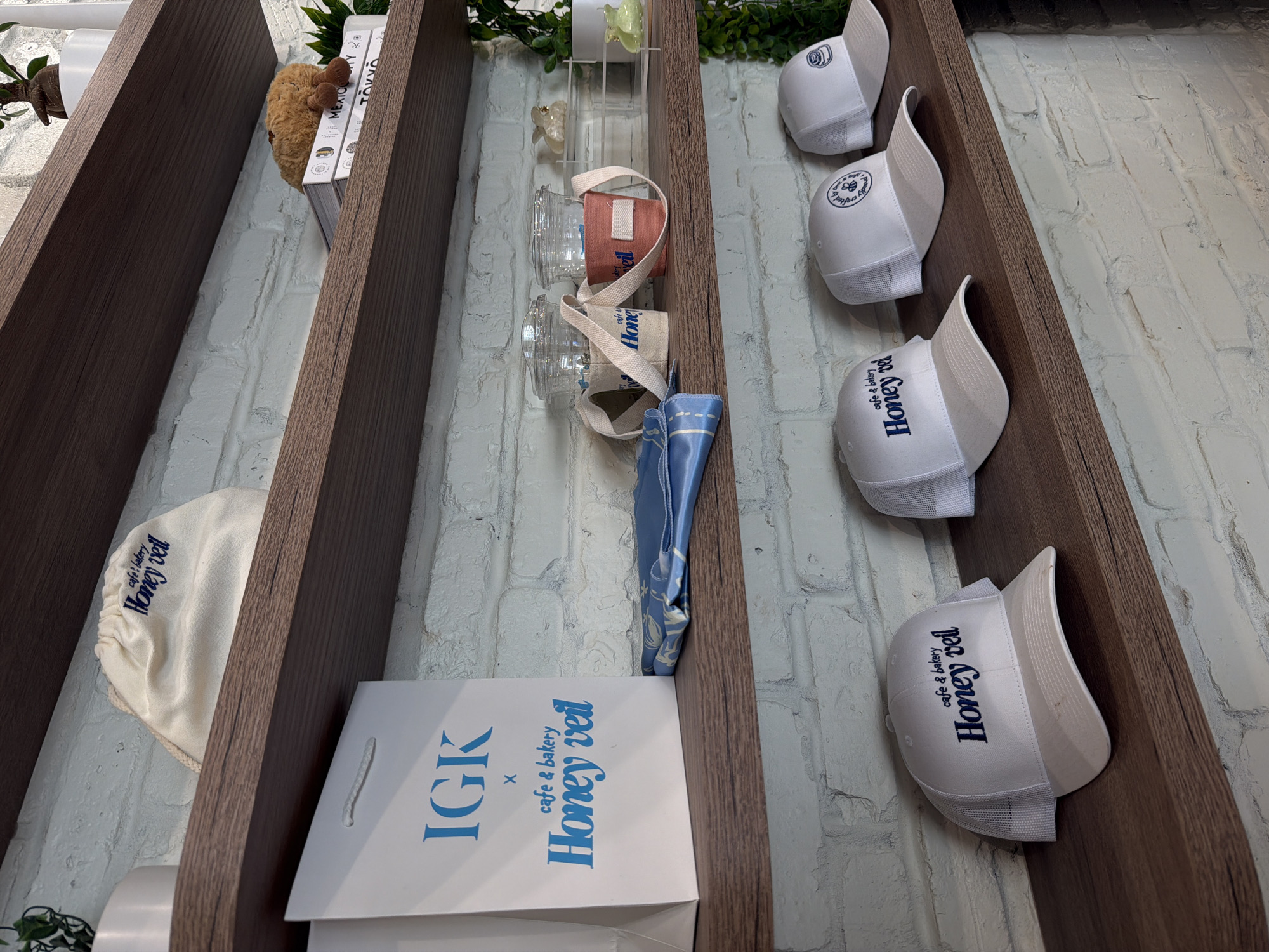 The image shows shelves in a shop displaying merchandise. On the top shelf, there is a white drawstring bag with "Honey veil cafe & bakery" printed in blue. To the right, a stack of books with a small stuffed animal on top.
The second shelf features a white paper shopping bag with blue text reading "IGK x cafe & bakery Honey veil". Next to it are two clear plastic cups with lids, each inside a fabric sleeve. One sleeve is beige and the other is coral. A folded blue cloth item is also on this shelf.
The bottom shelf displays several white mesh-backed baseball caps with blue embroidery. The caps have logos and text that appear to be related to "Honey veil cafe & bakery".
The background is a white brick wall. The setting is likely a retail store, possibly a cafe or boutique, located in South Miami, United States of America.