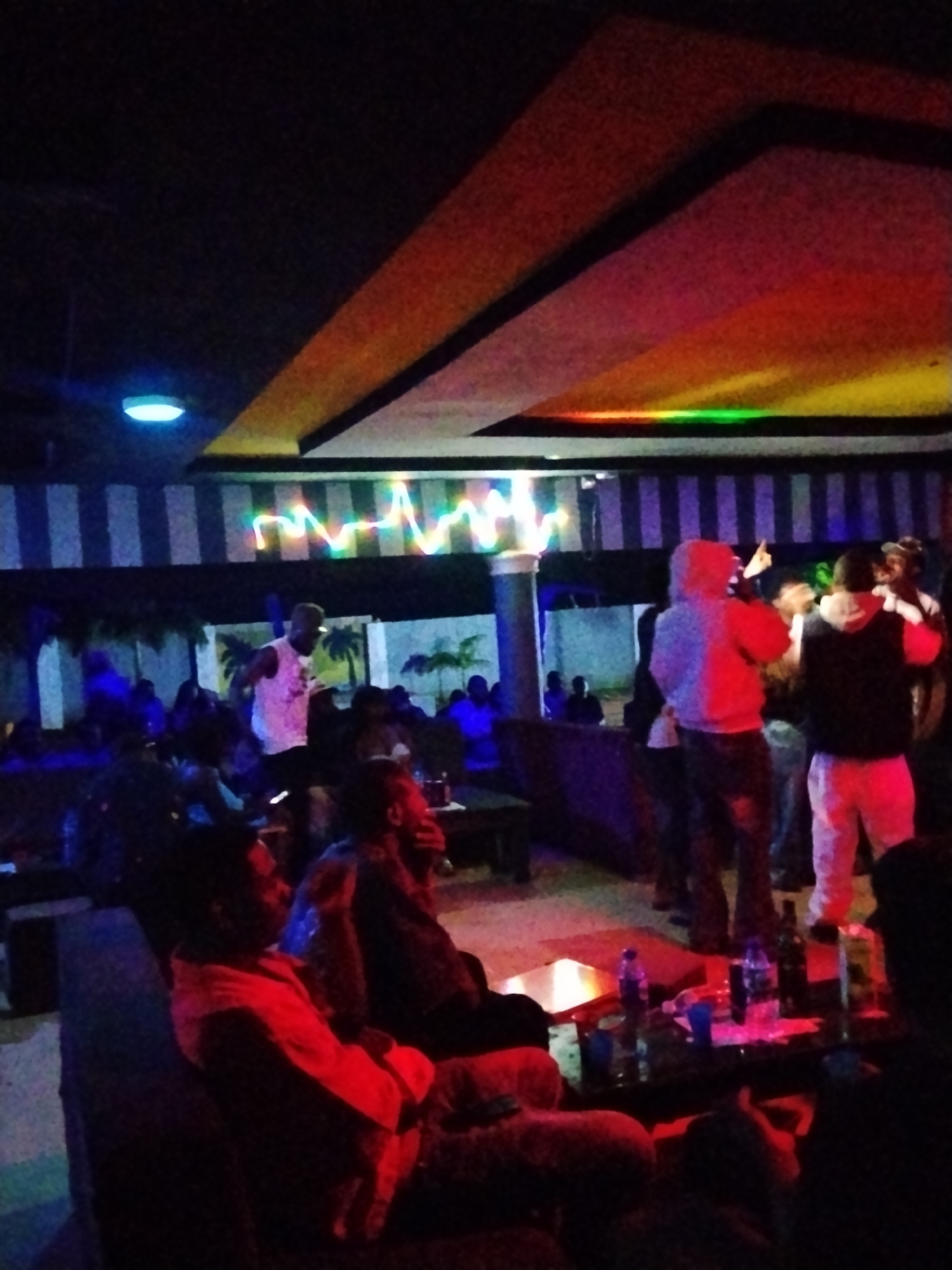 The image depicts a dimly lit indoor venue, possibly a club or bar, located in Durumi, Nigeria. The environment is characterized by colored lighting, with a striped ceiling and a multi-colored light fixture overhead. Several people are present, some standing and some seated.
In the foreground, a person is seated on a dark couch, illuminated by red light, with their body turned to the left. To their right, another person is partially visible, also seated.
In the mid-ground, several individuals are standing and interacting. One person, wearing a white tank top and dark pants, is performing or speaking in front of a crowd. To the right of this performer, three individuals are standing together. One is wearing a light-colored hoodie, another a dark hoodie and light-colored pants, and a third is largely obscured but appears to be wearing a dark shirt.
Further in the background, more people are seated and standing, their forms largely obscured by shadows and colored lighting. A table with bottles and cups is visible on the right side of the mid-ground. The overall atmosphere suggests a social gathering or entertainment event.