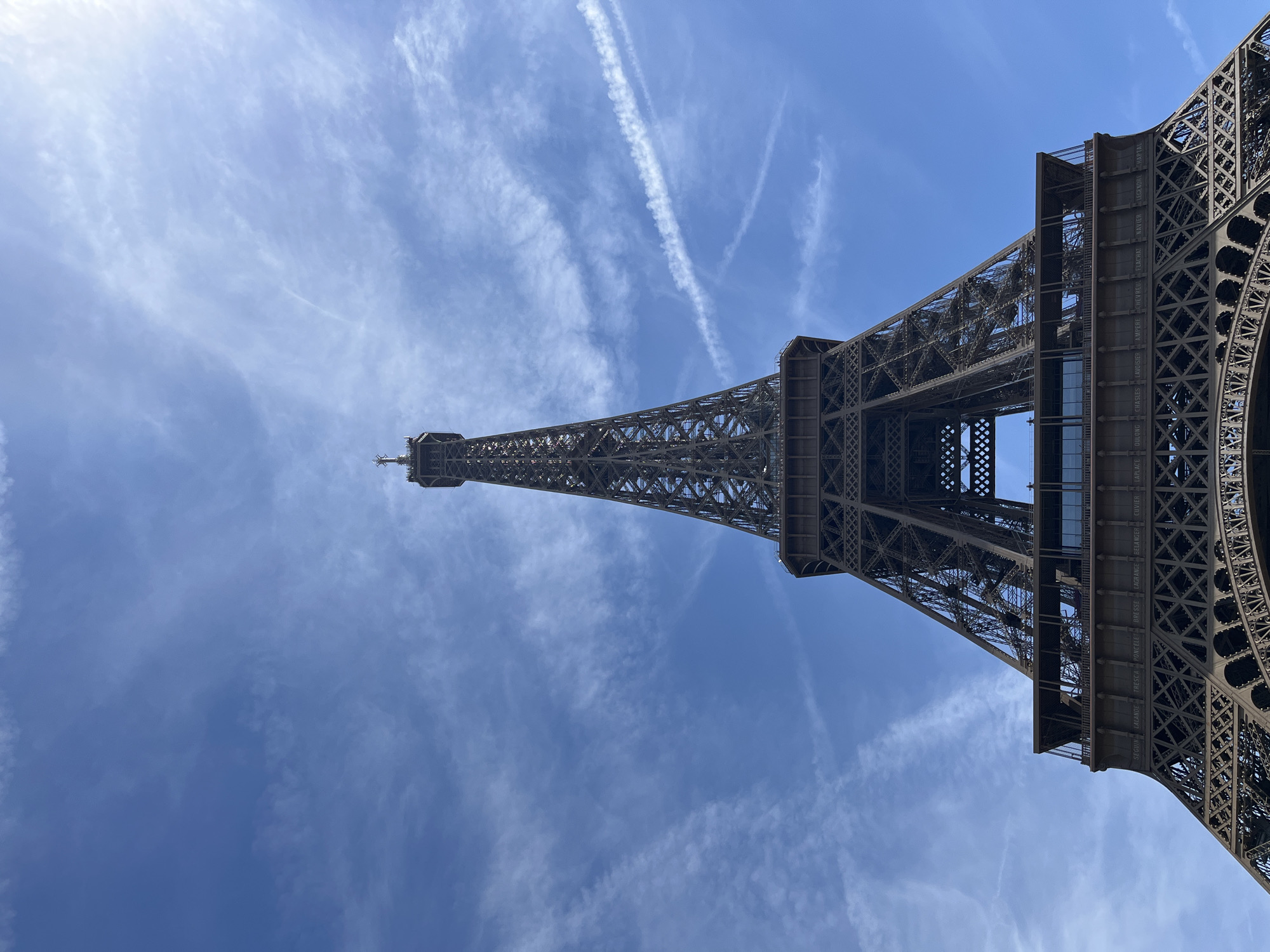 The image is a low-angle, full-shot photograph of the Eiffel Tower in Paris, France. The tower is the primary subject, dominating the center of the frame as it extends upwards towards a clear blue sky with wispy white clouds and contrails. The intricate iron lattice work of the tower is visible, with its iconic structure tapering to a point at the top. The bottom portion of the tower, including its base and the first level with its visible balconies and signage, is also present in the foreground. The sky occupies the upper two-thirds of the image, providing a bright and open backdrop. The lighting suggests it is daytime, with the sun likely positioned to the right and slightly above the viewer, casting a soft illumination on the tower. No people or other objects are clearly discernible in the image. The scene captures the grandeur and architectural detail of the Eiffel Tower against a serene sky.