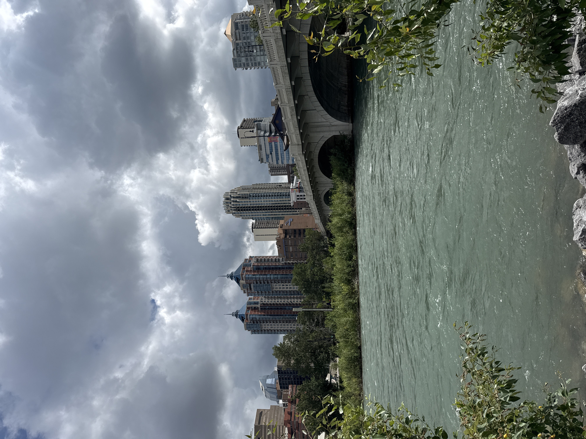 This image captures a scenic view of the Bow River in Calgary, Canada, with the city's downtown skyline as a backdrop. The river, a murky greenish-blue, flows with noticeable ripples, suggesting a steady current. In the foreground, leafy green branches frame the lower left and right corners of the image, with large gray rocks visible along the riverbank on the right.
Dominating the mid-ground is a multi-arched concrete bridge spanning the river, its stone railing casting shadows. Beyond the bridge, a dense line of green trees and bushes separates the river from the urban landscape. The city skyline features a cluster of modern high-rise buildings, some with distinctive architectural elements like pointed roofs on twin towers, and others with more contemporary glass facades. The buildings are varied in height and design, creating a dynamic urban silhouette.
The sky above is heavily overcast with dramatic, thick gray clouds that obscure any direct sunlight. Patches of lighter sky can be seen through breaks in the clouds, hinting at the possibility of sunshine elsewhere. The overall impression is of a slightly moody, overcast day in an urban setting adjacent to a significant natural waterway. No people are visible in the frame.
