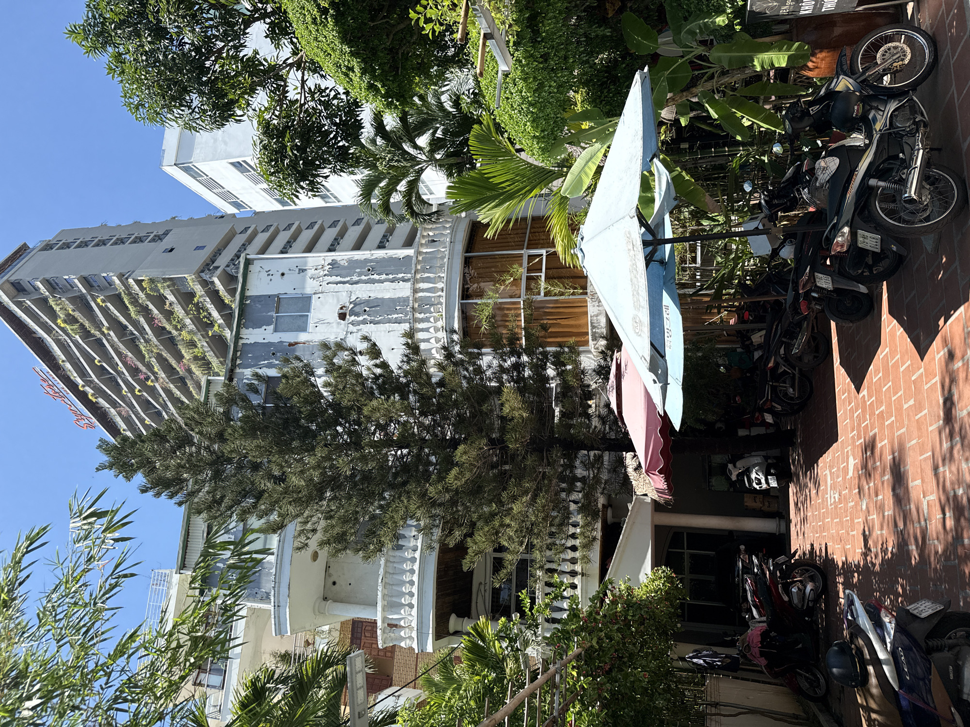 The image captures a bright, sunny day in Nha Trang, Vietnam. The scene is an outdoor area adjacent to a multi-story building with a faded, white facade and balconies. Lush green vegetation, including palm trees and leafy bushes, surrounds the building and spills onto the brick-paved ground. Several motorcycles are parked along the side of the building, suggesting a residential or commercial area. A large, light blue umbrella, partially open, stands in the foreground. The architecture of the building, with its repeated windows and decorative railings, hints at a tropical urban environment. Shadows from the trees and the umbrella are cast on the brickwork, indicating the time of day is likely midday. The overall impression is one of a warm, relaxed atmosphere in a bustling coastal city. Visible text on a motorcycle license plate reads "92H1-018.92".