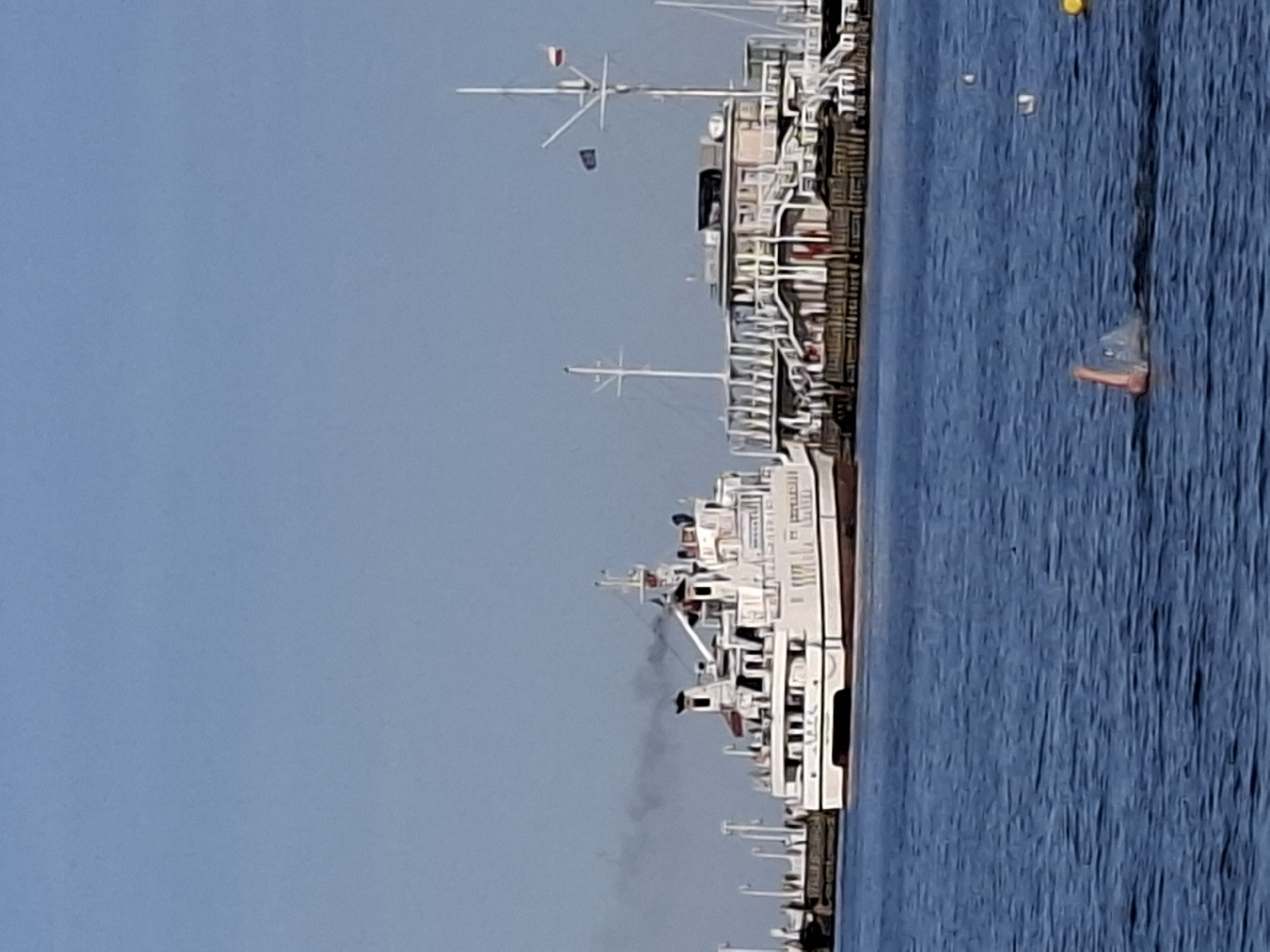 A white steamship is docked at a pier in Sopot, Poland. The steamship is emitting a plume of dark smoke from its smokestacks. Adjacent to the steamship is a multi-story building with white railings and a red roof. In the foreground, a person is swimming in the blue water, with an arm raised out of the water. The sky is clear and blue.