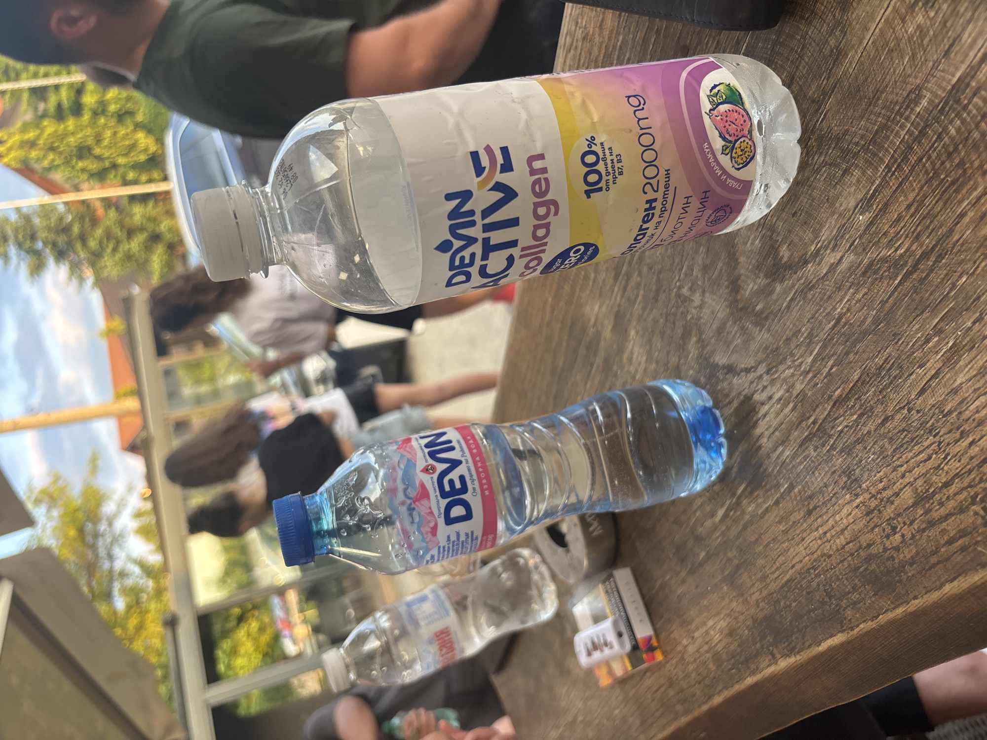 Here is a description of the image.
The image shows a wooden table surface with several items and people in the background, possibly in Plovdiv, Bulgaria.
On the table are two brands of bottled water, "Devin," and "Devin Active Collagen" with guava and passion fruit flavors. A package of what appears to be cigarettes or tobacco products is also visible.
In the background are several people in casual clothing, some sitting and others standing. There is a view of a building or structure with glass panels and possibly some greenery. The lighting indicates it's daytime.