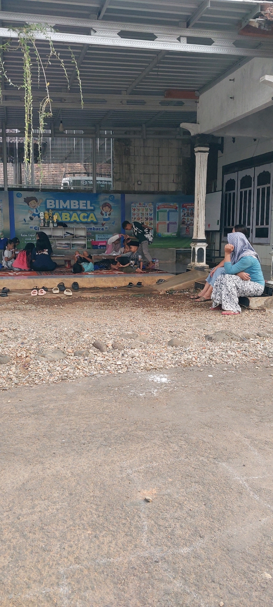 In the city of Keling, Indonesia, a group of children and adults are gathered in an outdoor educational setting. The scene takes place on a paved surface with scattered gravel and stones, in front of a building with a covered porch. A large mural adorns the back wall, featuring cartoon characters and the text "BIMBEL ABACA," indicating a reading tutorial center.
Several children are seated on mats on the ground, engaged in various activities. One child, wearing a black and white patterned shirt, appears to be interacting with another child who is wearing a black skullcap. Another group of younger children sits together, some looking at books. In the background, a few adult figures are present, including one woman in a blue headscarf and patterned pants, seated with her arm around another person. Another woman, also wearing a headscarf, is seated near the front, looking down and covering her face with her hand.
Various objects are scattered around the area, including pairs of flip-flops and sandals lined up near the edge of the paved area. Trophies are visible on a shelf behind the mural. The environment is somewhat informal, with a rustic outdoor feel, suggesting a community or local learning space. The building structure includes a corrugated metal roof supported by metal beams and a pillar on the right side. Sunlight filters through the covered area, illuminating parts of the scene.