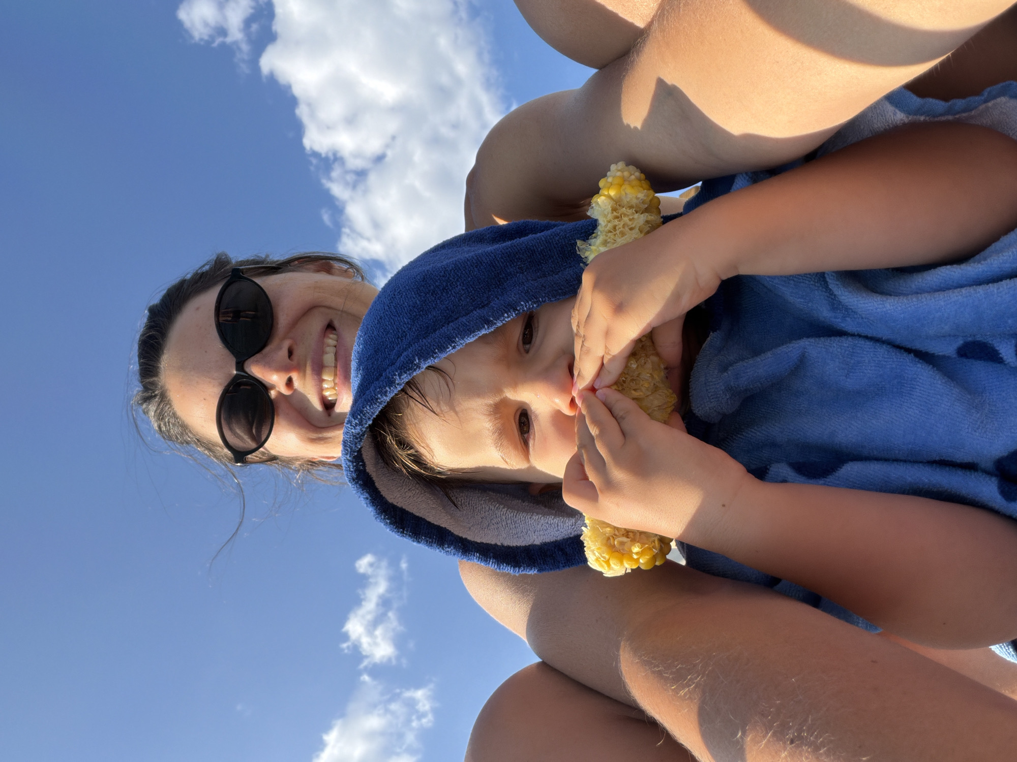 In Budva, Montenegro, a woman with sunglasses and a blue towel around her head is taking a selfie with a young child, who is also wrapped in a blue towel and eating corn on the cob. The sky is blue with some clouds. The image appears to have been taken outdoors during the day.