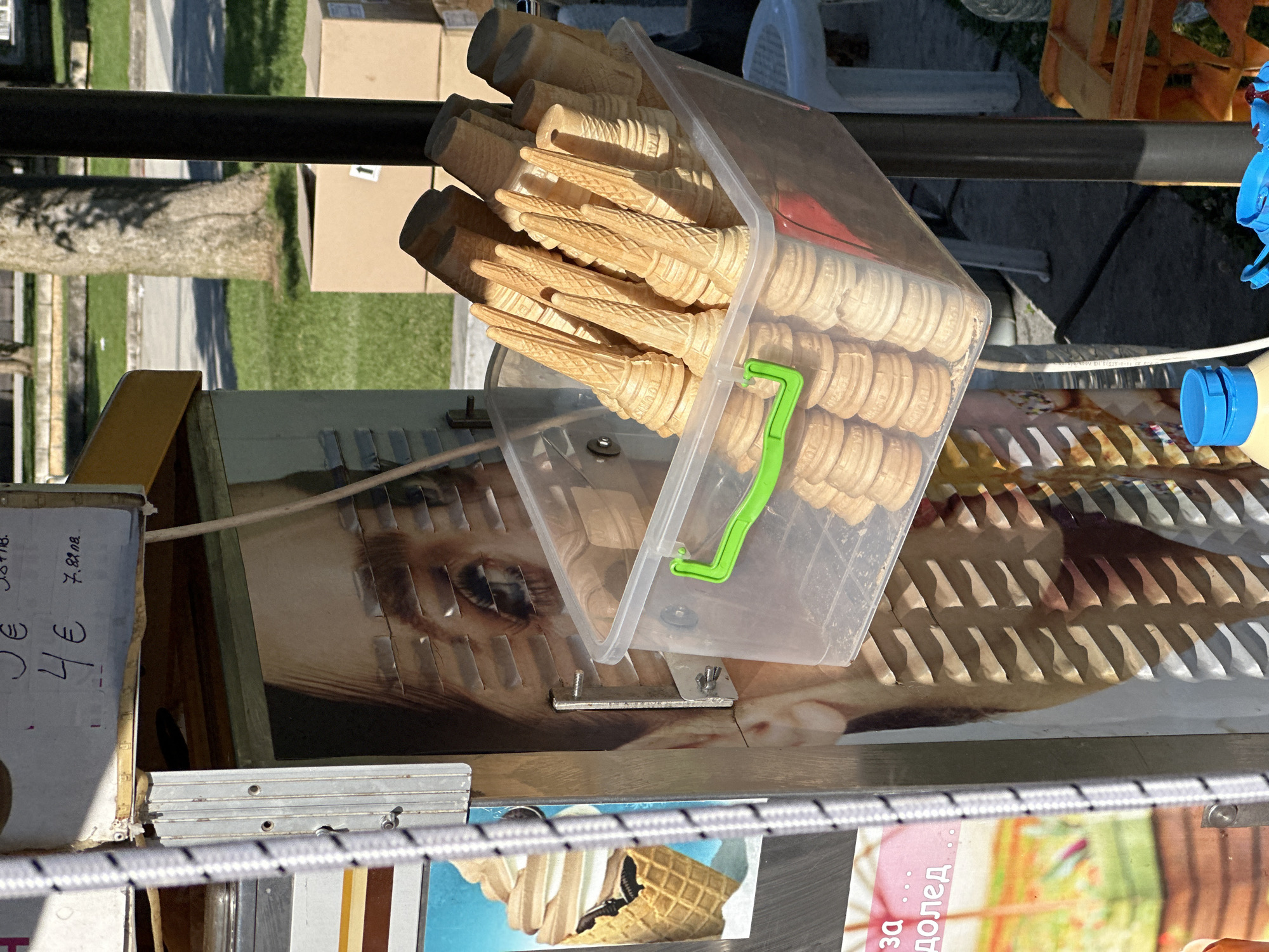 A close-up, angled shot shows a transparent plastic container filled with waffle ice cream cones. The container, secured with a green latch, rests on a surface displaying an image, partially obscured, of a human face. Price tags reading "4 €" and "7.80 лв." are visible. The photo was likely taken in Montana, Bulgaria, given the Cyrillic script seen on a lower poster showing images of ice cream. A black metal bar and other items are in the foreground.