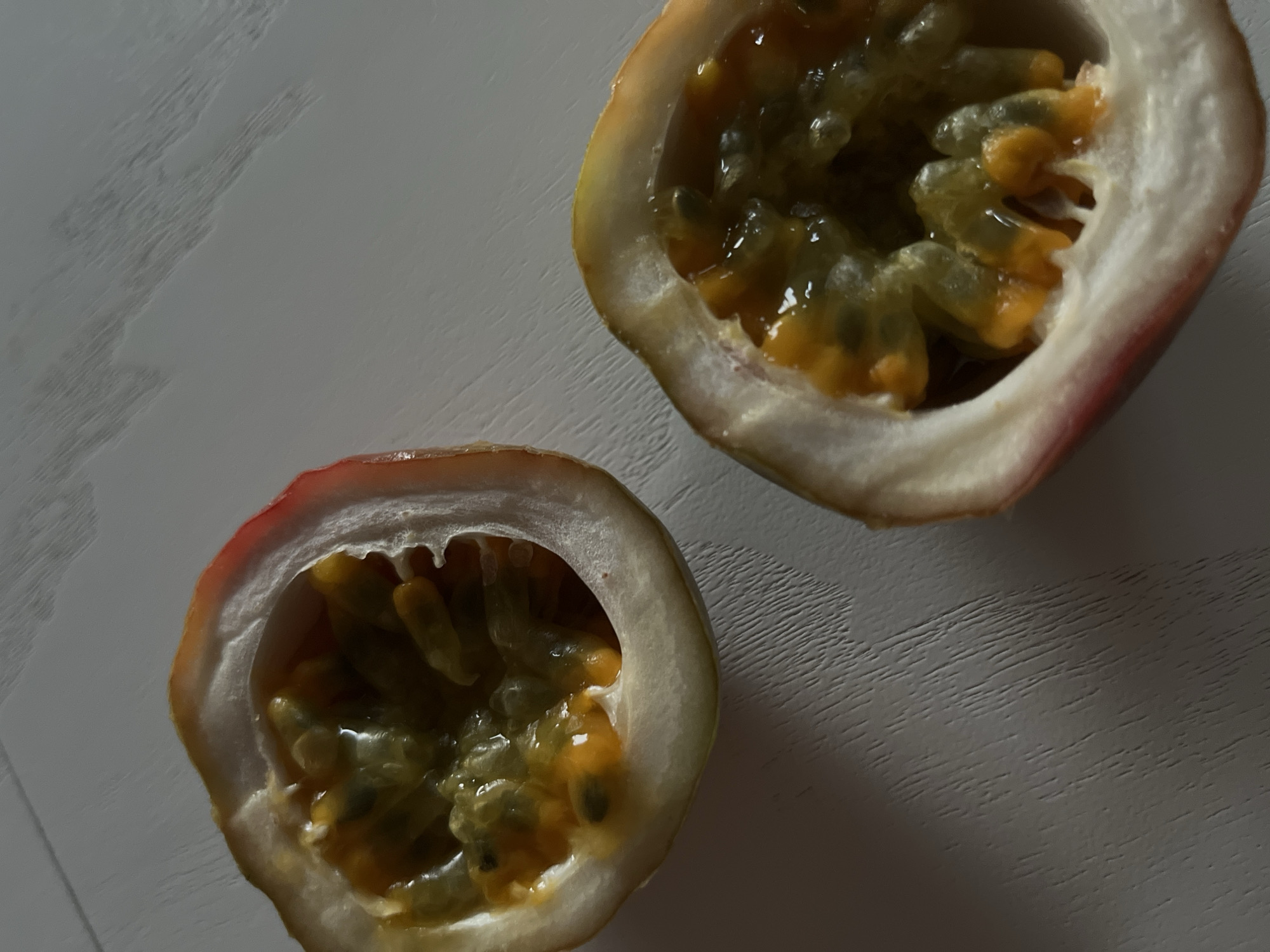 The image shows two halves of a passion fruit on a light-colored, textured surface. The fruit is cut horizontally, revealing its interior filled with seeds surrounded by jelly-like pulp. The pulp is a vibrant orange-yellow color, and the seeds are dark and numerous. The rind of the passion fruit is light yellow with a reddish-purple blush on the edges. One half of the passion fruit is positioned in the upper right of the frame, and the other is in the lower left. The surface the fruit rests on appears to be wood with a distinct grain pattern, possibly a table or countertop. The lighting suggests it is indoors, with a soft light source casting subtle shadows. There is no text visible in the image. The overall impression is a close-up, still-life shot of fresh fruit.