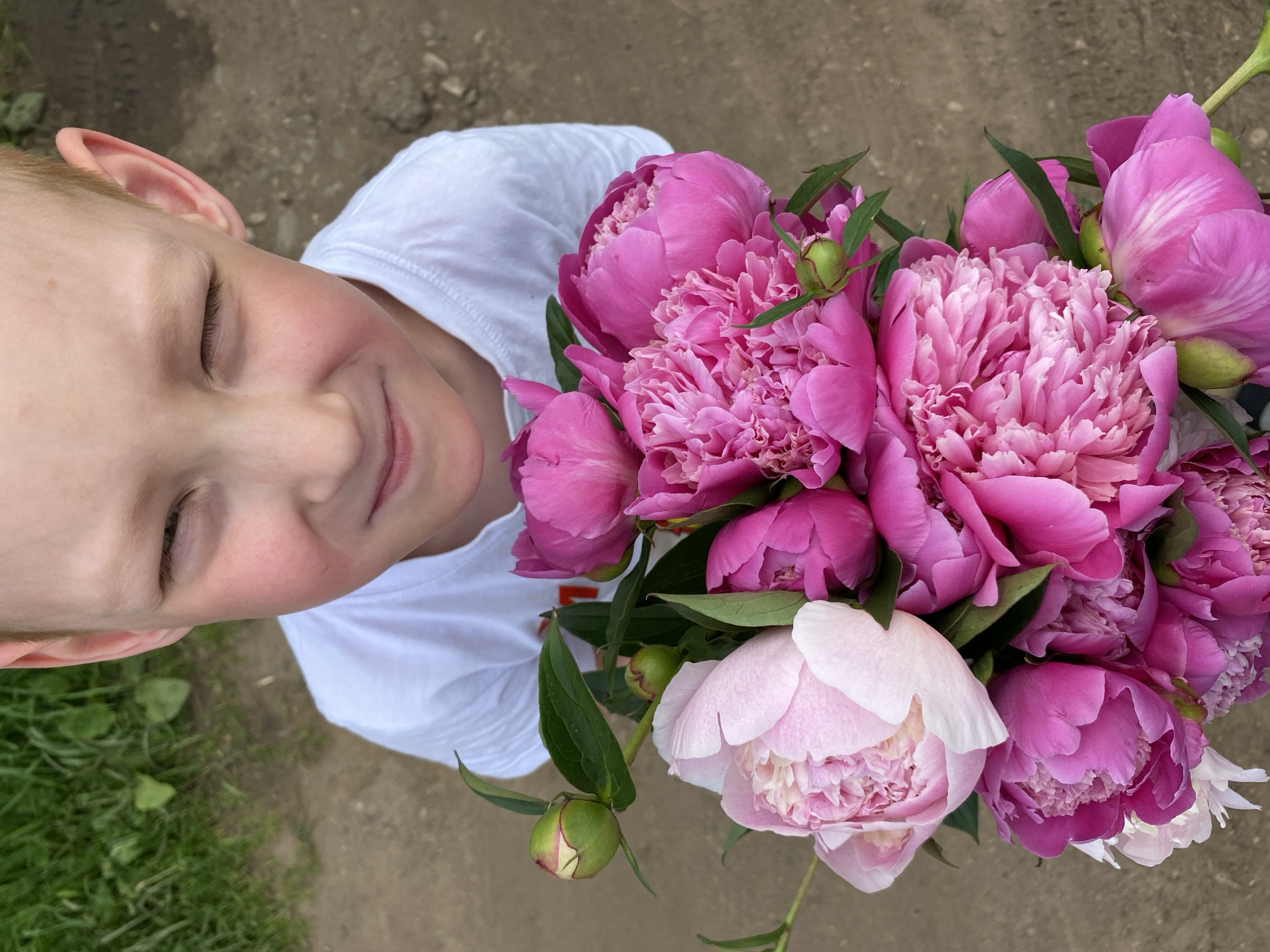 A young boy with light skin and reddish-blonde hair, wearing a white t-shirt, is looking down and to his right, with his right eye squinted. He is holding a bouquet of pink and light pink peonies. The background consists of green grass and dirt ground. The image appears to be taken outdoors in a park or garden area, possibly in the city of Chaikovsky, Russia, given the context.