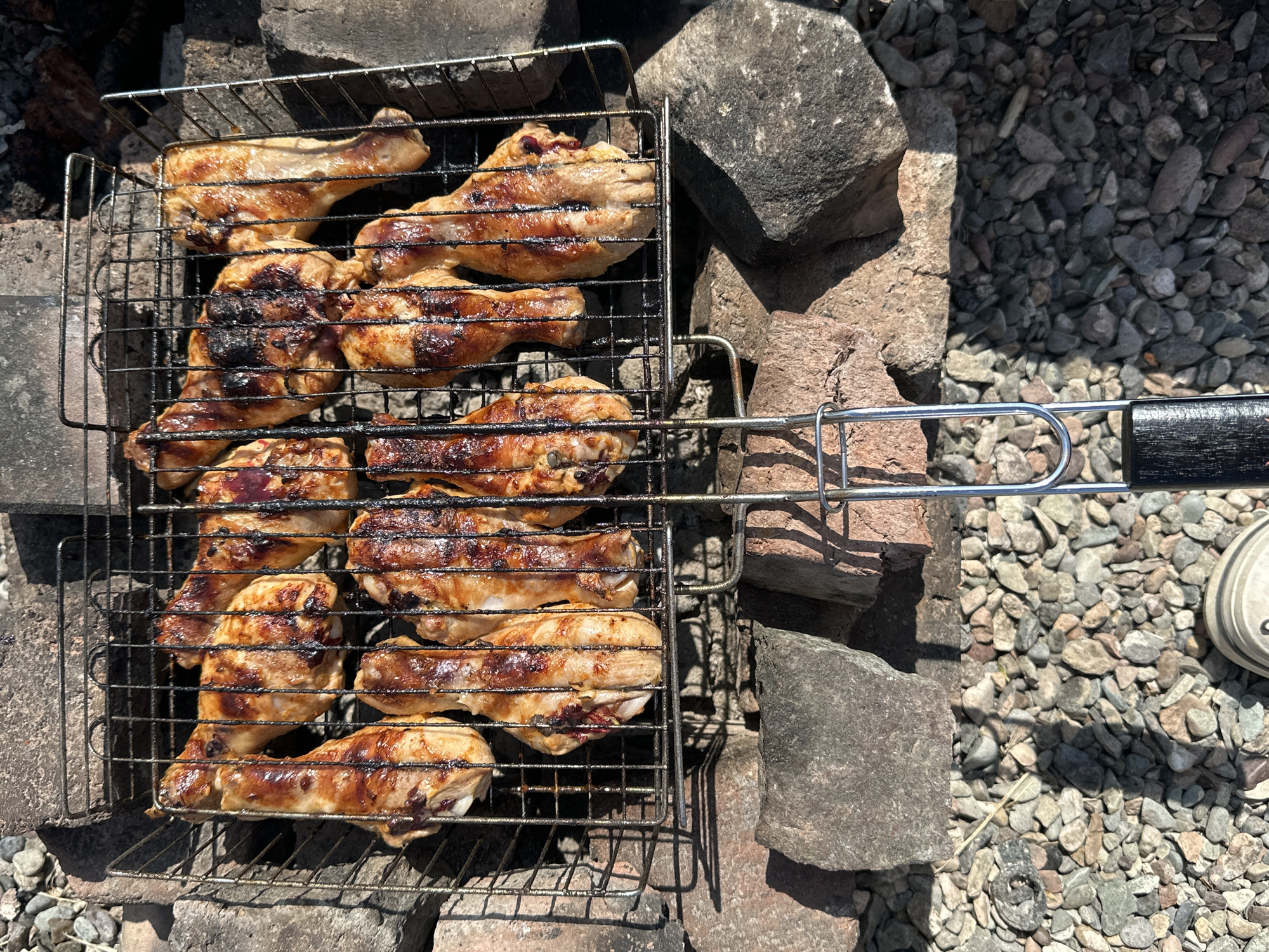 A barbecue grill basket filled with grilled chicken drumsticks is positioned over a makeshift fire pit constructed from stones and bricks. The chicken pieces exhibit char marks and a glistening, cooked appearance. The surrounding area consists of small stones and larger rocks, with some bricks visible. The lighting suggests an outdoor setting with sunlight casting shadows. The image appears to be taken from a high angle, looking down at the grill. The location is identified as Novonikolaevka, Russia.