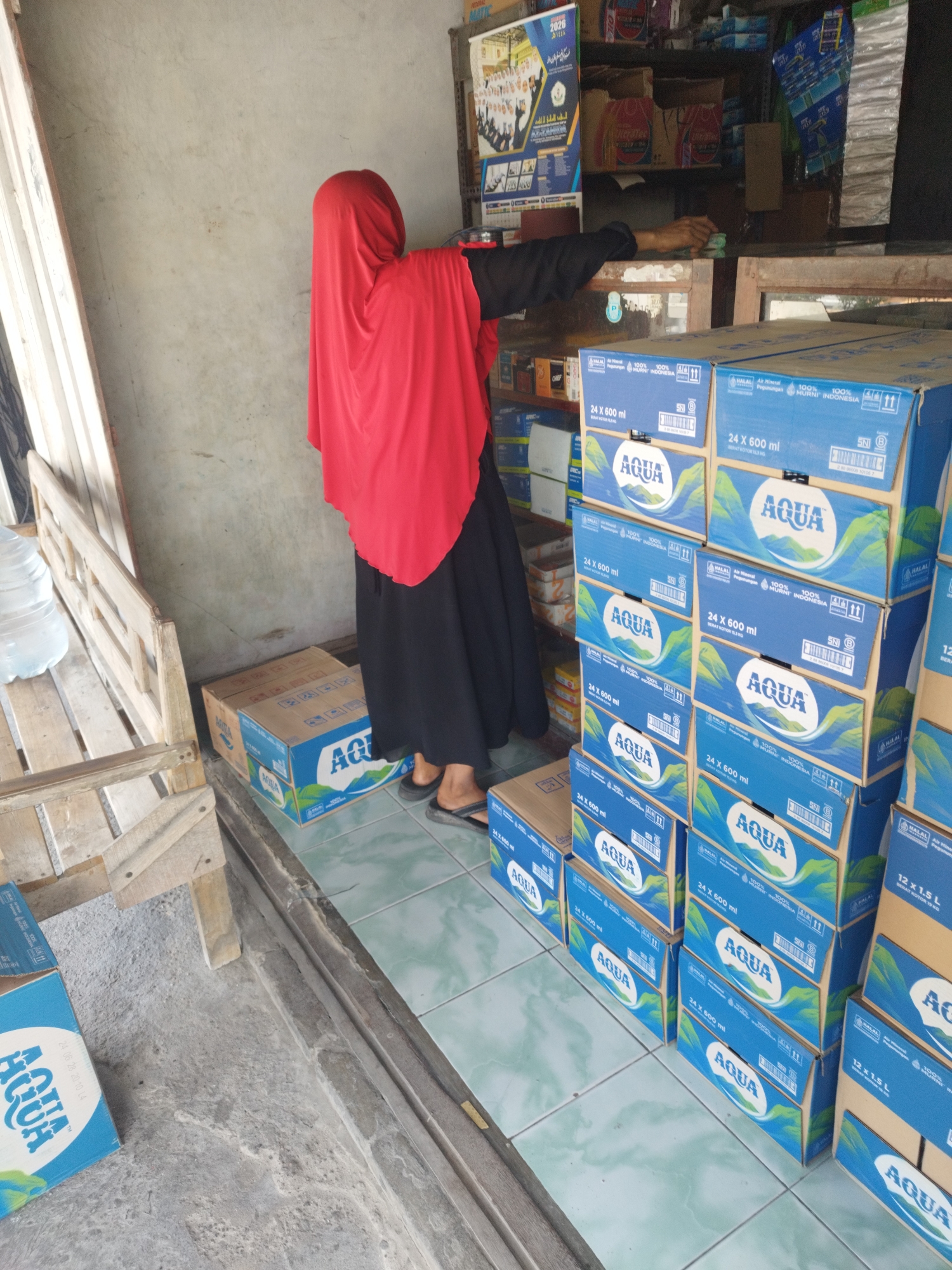 A person wearing a red hijab and a black abaya stands in front of shelves stocked with merchandise in a store. The person's right hand is extended towards the shelves, appearing to interact with items on a counter. In the foreground, stacks of blue cardboard boxes labeled "AQUA" are arranged on the floor. To the left, a wooden bench with a large water jug is visible. The environment suggests a small retail establishment, possibly a convenience store or minimarket, in the city of Woro, Indonesia. The walls are bare concrete, and the floor is tiled.