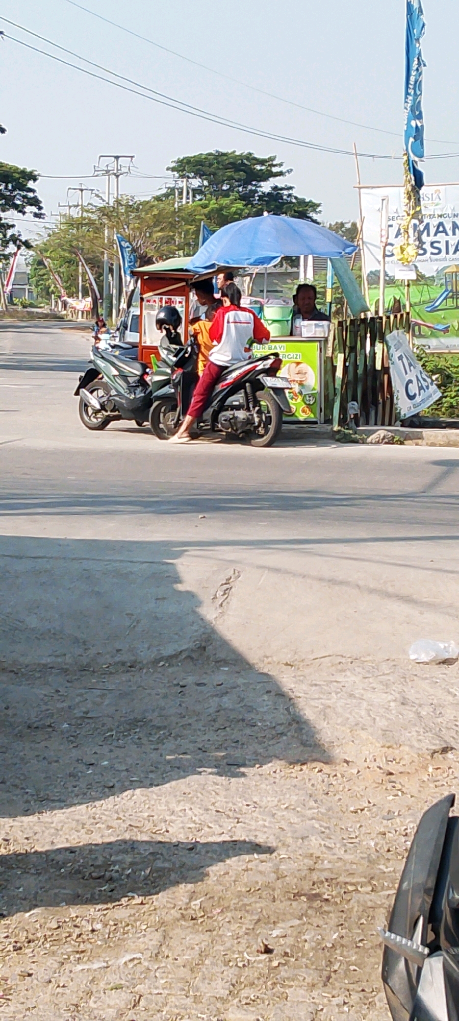 The image depicts a street scene in Sriamur, Indonesia, with a focus on a food stall under a blue tarp. Two individuals, likely customers, are seen near the stall. One is seated on a black motorcycle, wearing a red and white shirt and dark pants, while the other stands behind them, partially obscured. Another person, presumed to be the vendor, is visible behind the stall's counter, serving or interacting with the customers.
The food stall itself is a mobile cart with a red and white facade, displaying various items, though they are not clearly identifiable. Adjacent to the stall is a fence made of bamboo-like material, and a large white banner with text, partially visible, hangs on it. A blue flag with yellow accents is also prominent, tied to a tall pole.
Two motorcycles are parked near the stall. The one in the foreground, where the seated individual is, is black. Another motorcycle, also black, is parked to its left. The environment suggests a roadside setting with a paved road and a sidewalk or shoulder area. Trees and utility poles are visible in the background, indicating an urban or semi-urban environment. The lighting suggests it is daytime, with shadows cast on the ground.
