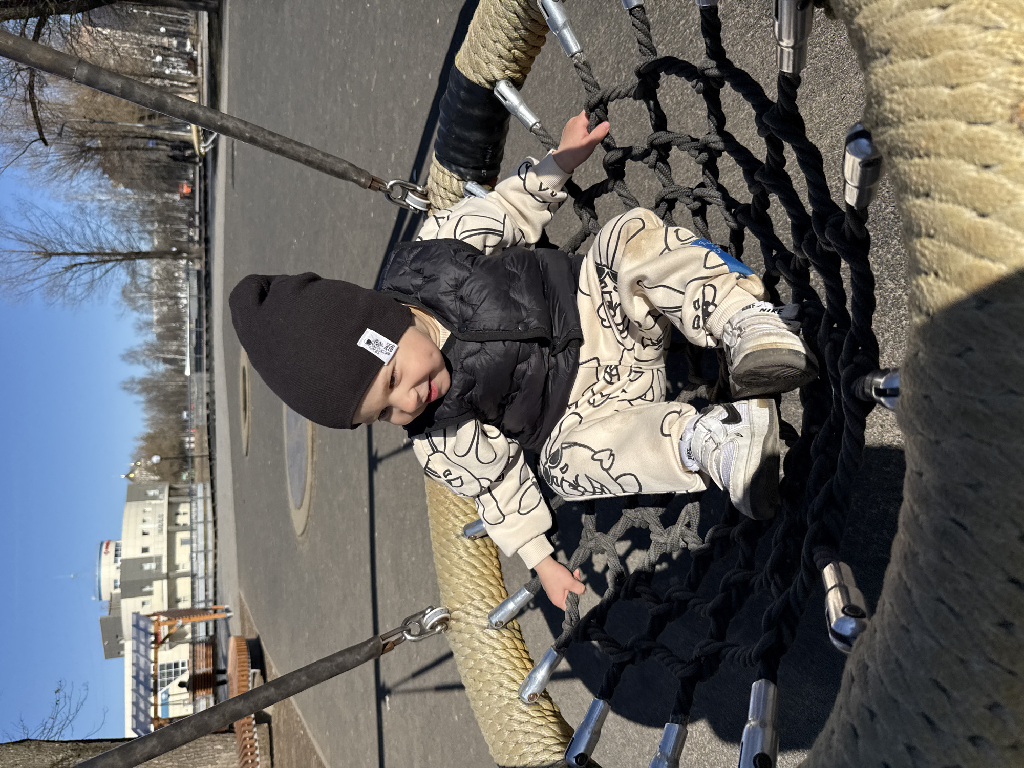 A young child wearing a black beanie, a black vest, and patterned pants sits on a large rope and net structure. The structure appears to be part of a playground. The child is holding onto the rope. In the background, there is a paved area, trees, and a building. The location is not specified in the details and it is not possible to provide city details.