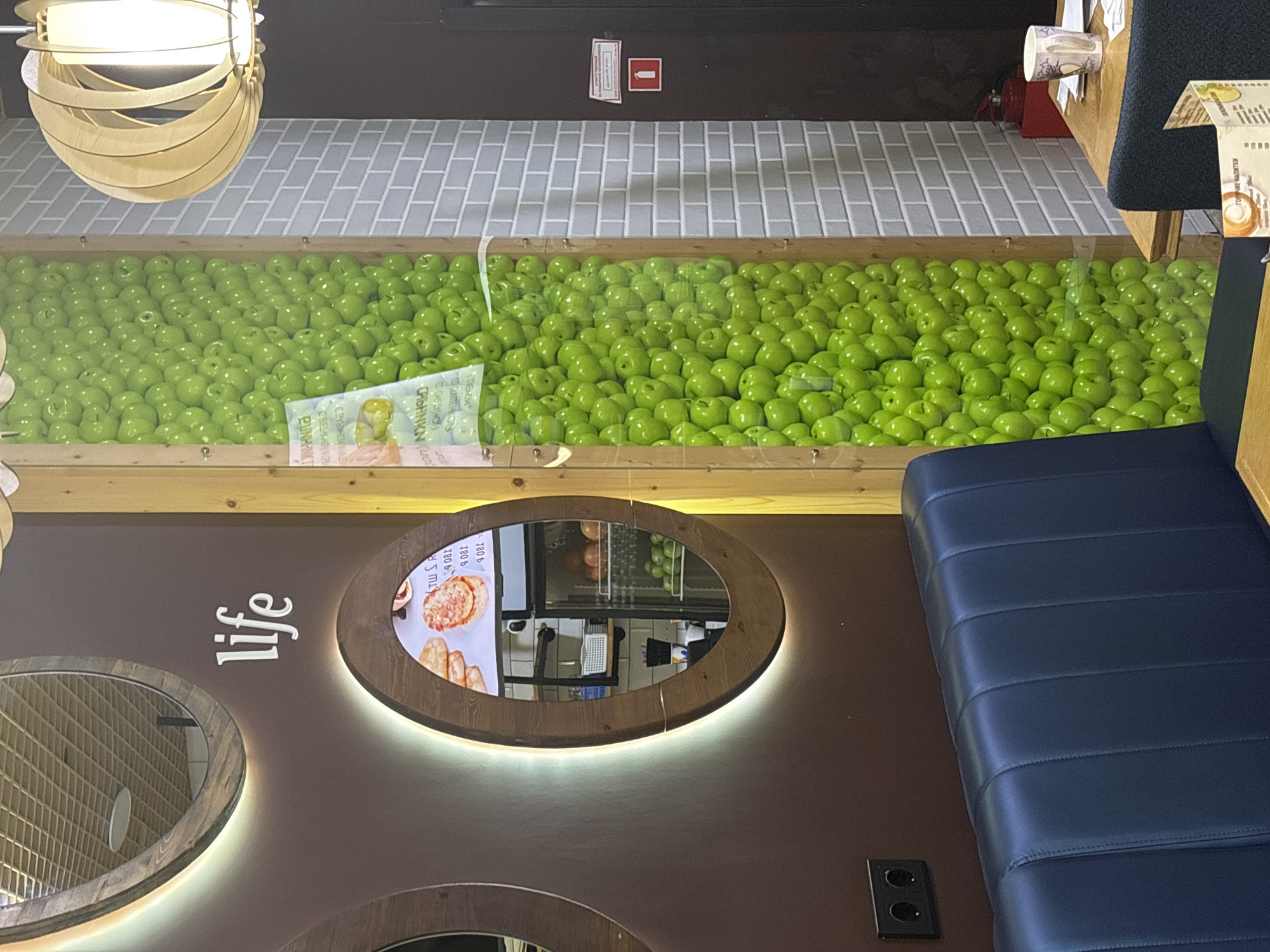 The image shows the interior of a cafe or restaurant. In the foreground, a dark blue upholstered bench is visible. To the left, a brown wall has a large, illuminated oval mirror. Above this mirror, the word "life" is displayed in white, stylized lettering. Further to the right, a tall, narrow wooden frame filled with numerous bright green artificial apples runs vertically. A light fixture with a wooden, slatted shade hangs from the ceiling to the right of the apple display. The background features a gray brick wall and a dark door. On a wooden table to the right, a mug and some papers are visible. The setting appears to be a modern, possibly themed establishment. The image does not contain enough information to determine the specific location in Naberezhnye Chelny, Russia.