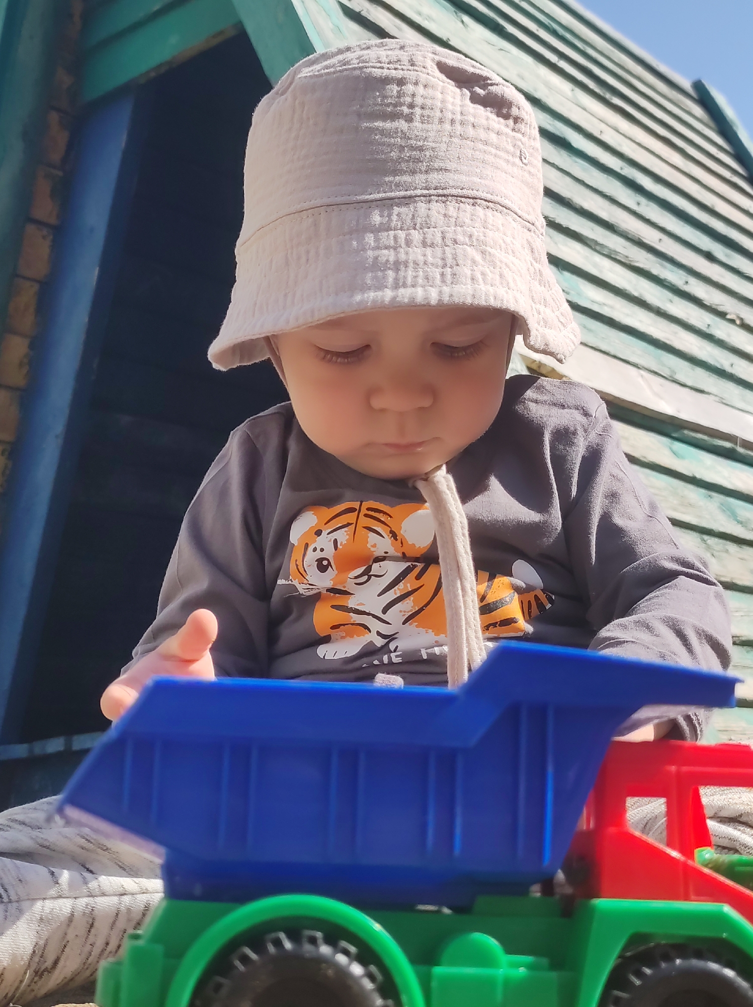 A young child wearing a light-colored bucket hat and a grey long-sleeved shirt with a tiger print is playing with a toy dump truck that is predominantly blue, red, and green. The child is seated in what appears to be a wooden play structure with green and blue painted planks. The location is likely outdoors, possibly in Chaikovsky, Russia, based on the request. The child appears focused on the toy, looking down at it. The scene is brightly lit.