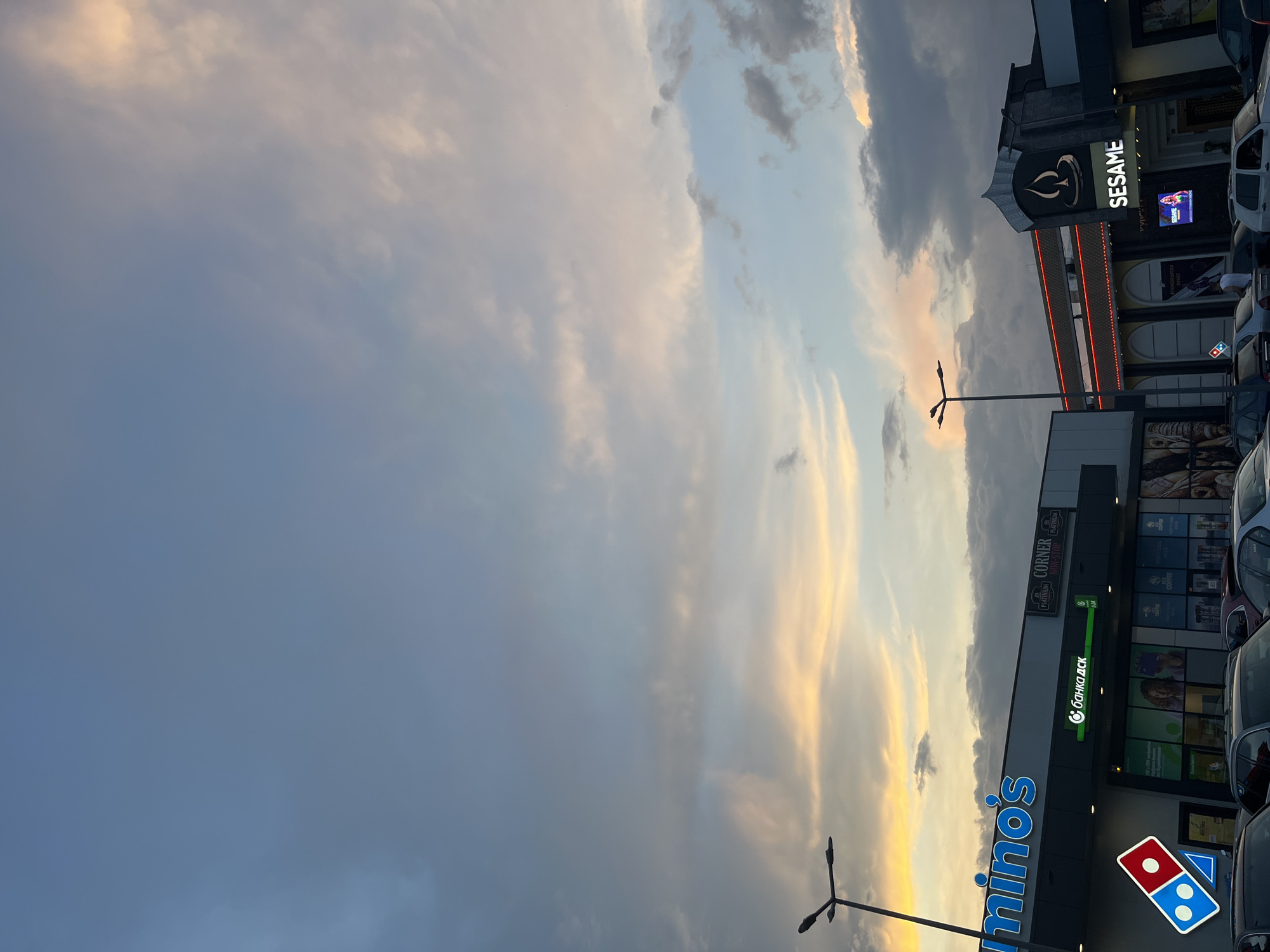 The image shows a sunset sky with dramatic clouds over a commercial street scene. A prominent blue sign with white lettering that reads "Domino's" is in the foreground on the left. To the right of the Domino's sign, a building facade features several businesses. One business has a sign with a stylized logo and the word "SESAME" in white lettering. Another business has a green sign that says "С банка дск" (which translates to "DSK Bank"). Above "DSK Bank," there is a sign for "CORNER" with "PLATINIUM" written above it. Cars are parked along the street in front of the buildings. The lighting suggests it is either early morning or late evening, with the warm glow of the sunset illuminating the clouds. The overall scene conveys a sense of everyday urban life at dusk, with a focus on commercial establishments and the sky.