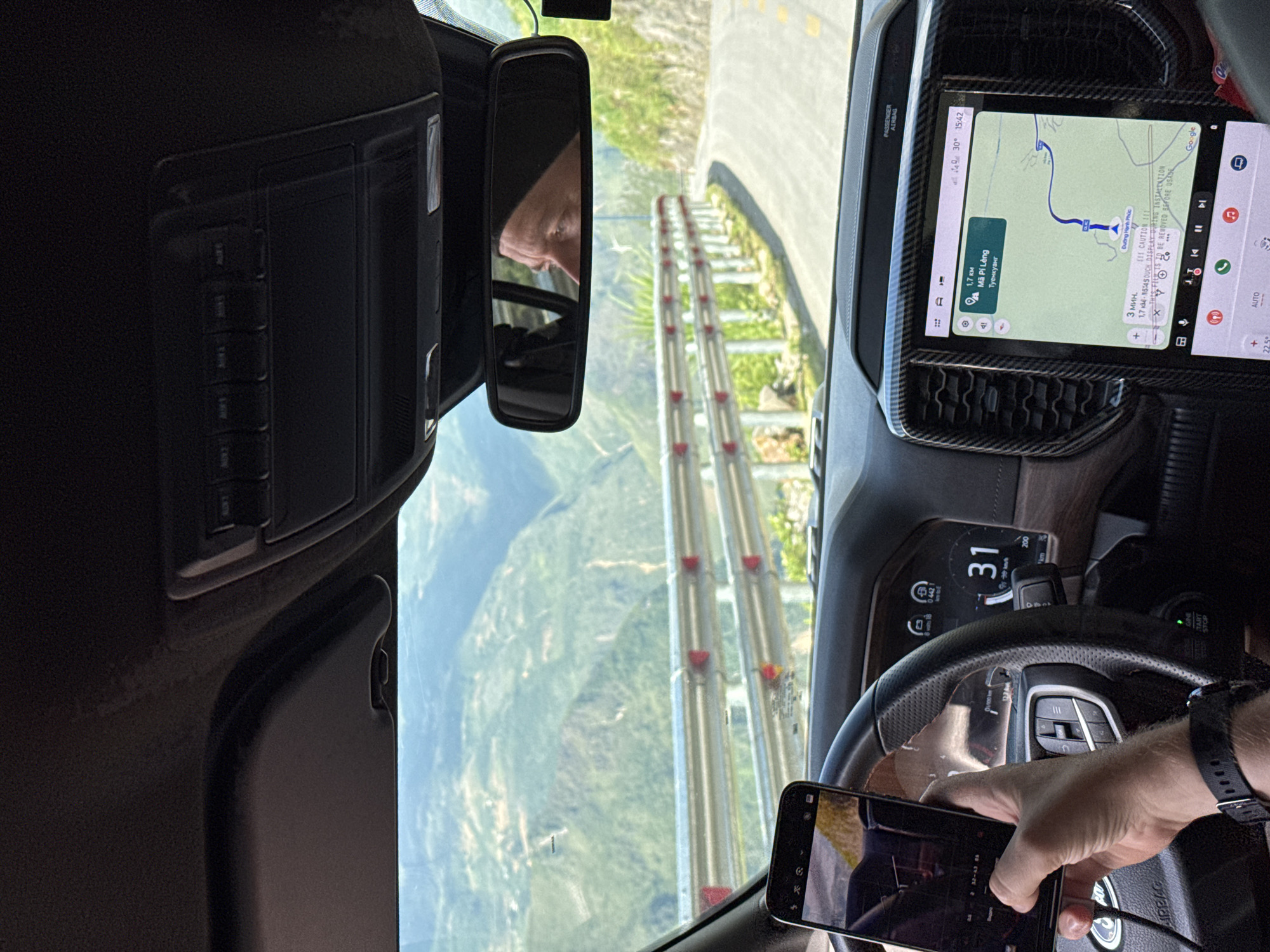 The image, taken from inside a vehicle, captures a view of a winding road with guardrails in Đồng Văn Commune, Vietnam. The navigation screen indicates the location is near Mã Pí Lèng, a mountain pass in the region, with a destination set for Dương Hạnh Phúc. The dashboard displays a speed of 31 km/h. The driver is visible in the rearview mirror and is holding a smartphone. The environment is mountainous and forested, typical of the region.