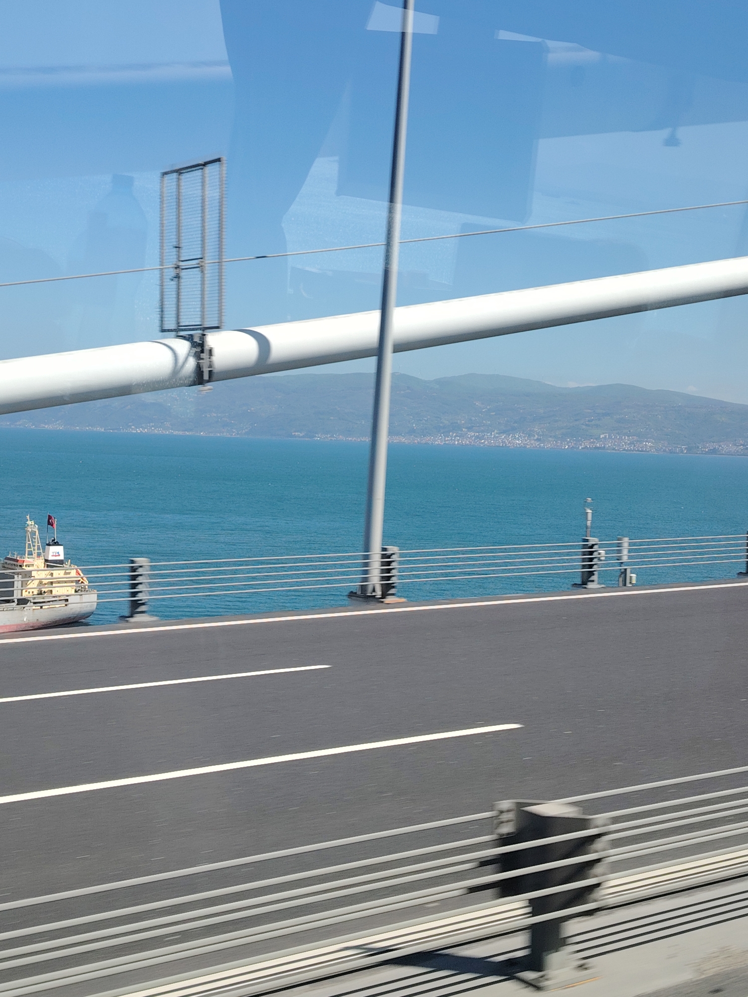 The image presents a view from inside a vehicle traversing a modern bridge over a vast body of water, likely the Gulf of İzmit near Dilovası, Turkey. The scene is brightly lit by daylight under a clear blue sky, with visible shadows indicating a sunny day.
In the foreground, the asphalt road of the bridge is visible with clear white lane markings. A sturdy metal railing system, consisting of multiple horizontal white bars and vertical supports, runs along the bridge's edge. Reflections, possibly of a person and the vehicle's interior, are visible on the window through which the photo was taken, especially in the upper left.
Spanning across the mid-ground is a large, white, cylindrical pipe-like structure, part of the bridge's support or cable system. A rectangular metal grid, possibly an antenna or sensor, is attached to this pipe, and a grey vertical pole extends upwards from the bridge deck.
Below the bridge level, the deep blue water of the gulf stretches towards a distant mountainous coastline. Settlements with numerous buildings are visible along the base of these mountains. On the left side of the water, a cargo ship or similar vessel is partially visible beneath the bridge. It features a grey hull, a white superstructure, and flies what appears to be a Turkish flag and another red flag from its mast. The letters "TPS" are discernible on the ship's side.
The overall impression is one of travel and impressive infrastructure set against a scenic natural backdrop.