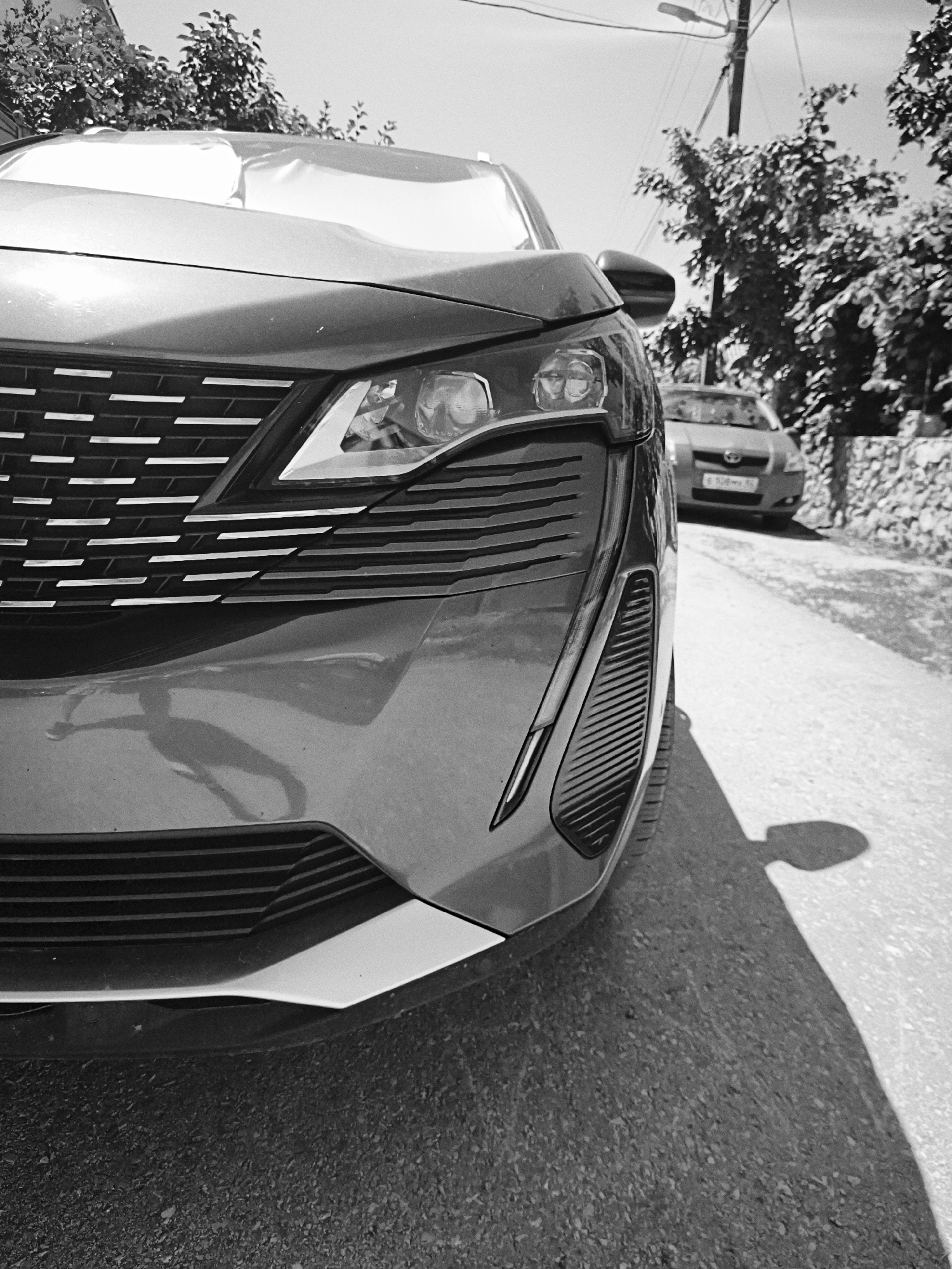 The image presents a detailed, low-angle, black-and-white view of a street scene located in Терновский округ, Ukraine, during daylight hours. A dark-toned, modern crossover or SUV, specifically a Peugeot 3008, occupies the left side of the foreground, with its front-right quarter prominently featured. The vehicle's complex grille with horizontal slat patterns and its distinctively shaped headlight cluster with integrated LED daytime running lights are clearly visible. A long, dark shadow, indicative of a person or object immediately to the left of the frame, stretches across the lower front bumper of the foreground vehicle and onto the textured asphalt road surface.
The asphalt road recedes into the background, where a lighter-toned sedan, identifiable as a Toyota Yaris, is visible further down the street, its front facing away from the camera. A partial view of its license plate, "E 108 MB 93," is discernible. To the right of the road, a low, rugged stone wall runs along the pavement, behind which a cluster of dense-foliaged trees grows. Additional trees are visible in the upper left and upper right background. A utility pole, equipped with a streetlamp, stands in the upper center-right portion of the frame. The sky above is bright and clear. No individuals are directly visible within the scene, and no explicit actions or interactions are occurring.