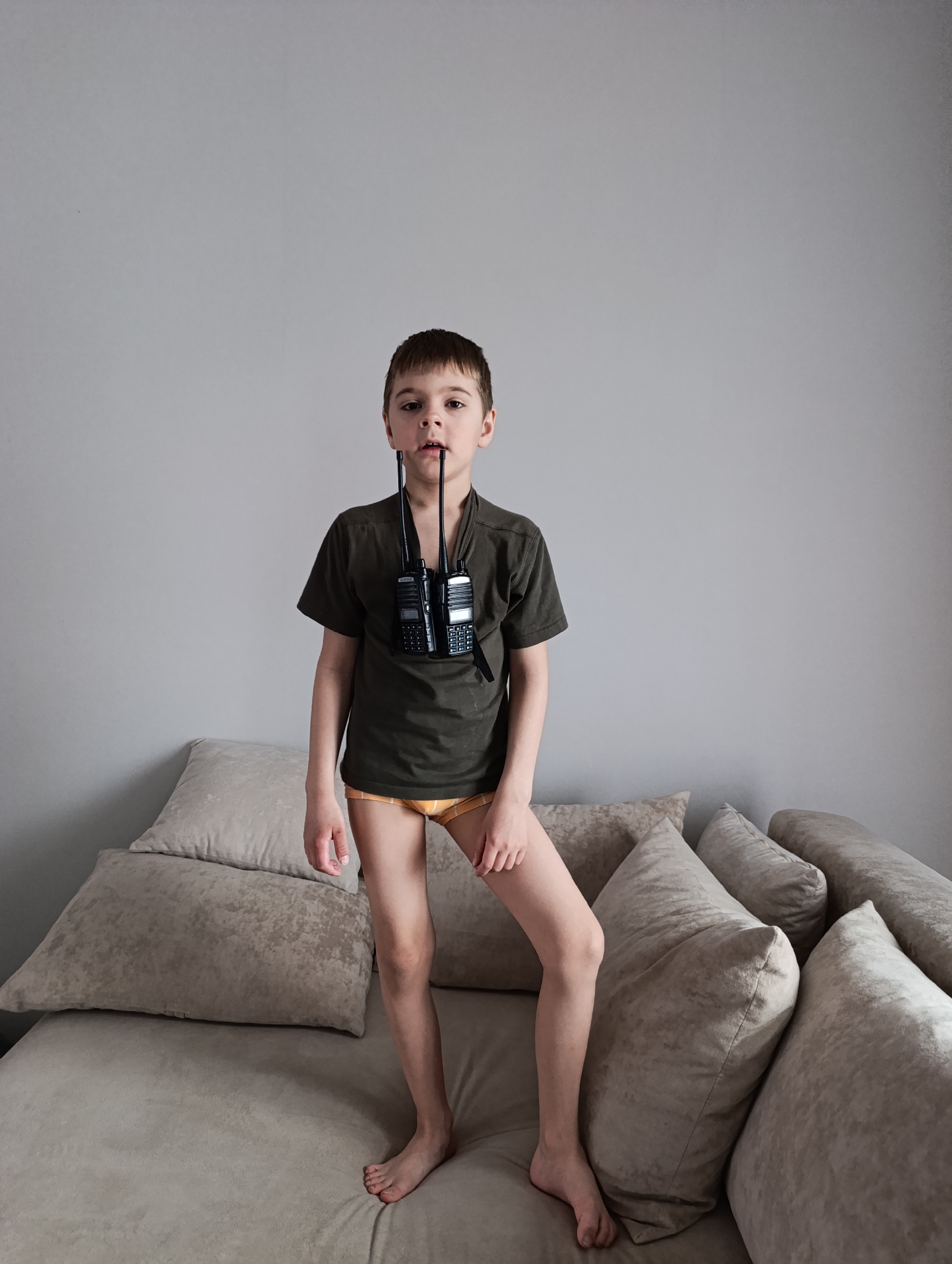 In an indoor setting, likely a home in Chaikovsky, Russia, a young boy is captured standing on a light-colored sofa or bed. He is barefoot and appears to be of elementary school age, with short brown hair. He wears a dark olive green short-sleeved t-shirt and light yellow and orange striped underwear.
Hanging around his neck are two black walkie-talkies, or two-way radios, positioned in front of his chest. Their antennas extend upwards, reaching past his chin. The boy looks directly at the camera with a neutral expression, his mouth slightly ajar.
The background consists of a plain light grey wall, suggesting a simple interior space. The sofa or bed he stands on is covered with several large, plush pillows in varying shades of beige or light grey, creating a soft and somewhat informal environment. The lighting suggests it is daytime. There is small, unreadable text visible on the bodies of the walkie-talkies.