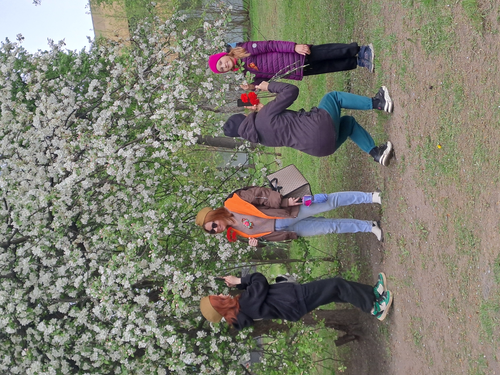 The image captures a park scene in Elektrostal, Russia, featuring four individuals standing near a blossoming tree. From bottom to top, there is a person with red hair wearing a brown cap and black attire with green shoes, then an adult in a brown jacket with an orange shirt and sunglasses holding red flowers, above them is a person wearing a black hooded coat holding the same type of flowers, and finally a child with a pink hat in a purple jacket. The blossoming tree with white flowers serves as a backdrop. The ground is covered in grass with some patches of dirt.