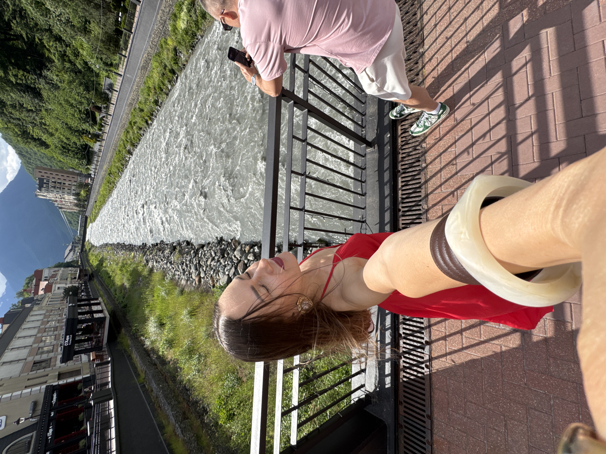 In the city of Estosadok, Russia, a woman in a red dress is taking a selfie on a bridge with a river running beside it. She is wearing large gold earrings and her hair is blowing in the wind. To her right, a man in a pink t-shirt and white shorts is leaning on the bridge railing, looking at his phone. The river is wide and turbulent, with rocky banks. On the left side of the river, there are buildings and trees, and on the right side, there is a road and a densely forested hill. The bridge has a metal railing and a tiled walkway. Shadows from the railing are cast on the walkway.