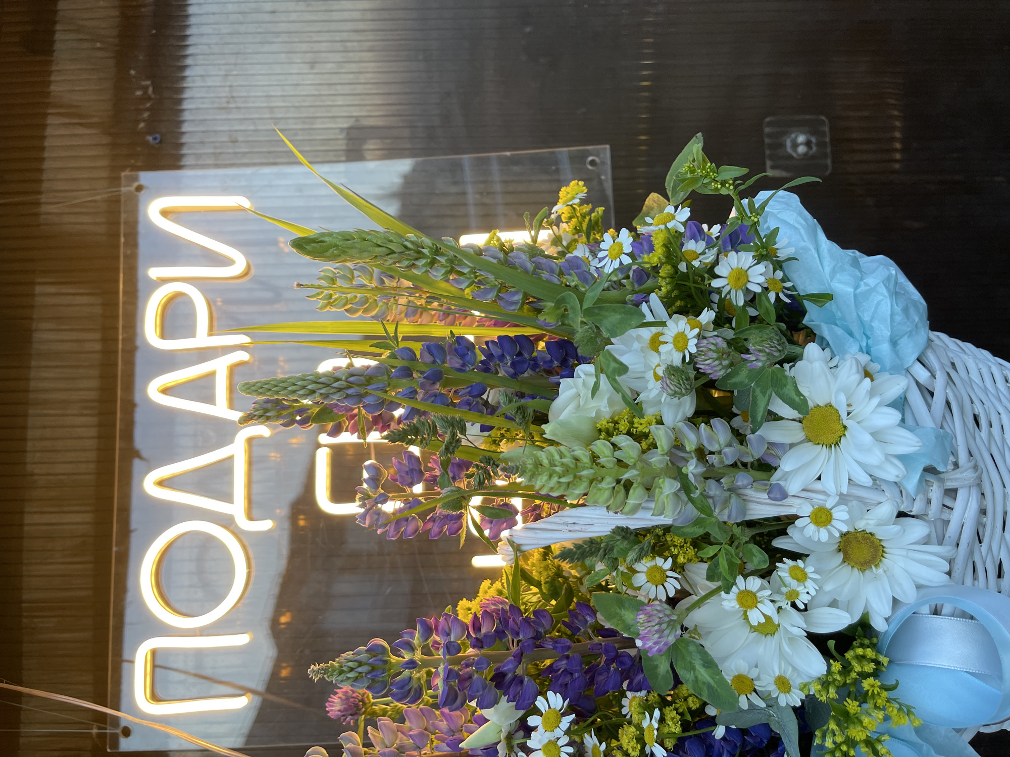 A white wicker basket overflows with a vibrant bouquet of flowers, including purple lupine, white daisies, and small yellow blossoms, with a light blue tissue paper lining the basket. Behind the flowers, a neon sign glows with Russian lettering that reads "ПОДАРИ" (meaning "GIFT"). The sign is mounted on a clear acrylic panel and is illuminated with a warm yellow light. The background appears to be a dark, ribbed surface, possibly a corrugated wall or siding. The lighting suggests it is either dawn or dusk, with the warm glow of the neon and the setting sun casting long shadows. The overall scene suggests a flower shop or a storefront in the city of Kommunarka, Russia.