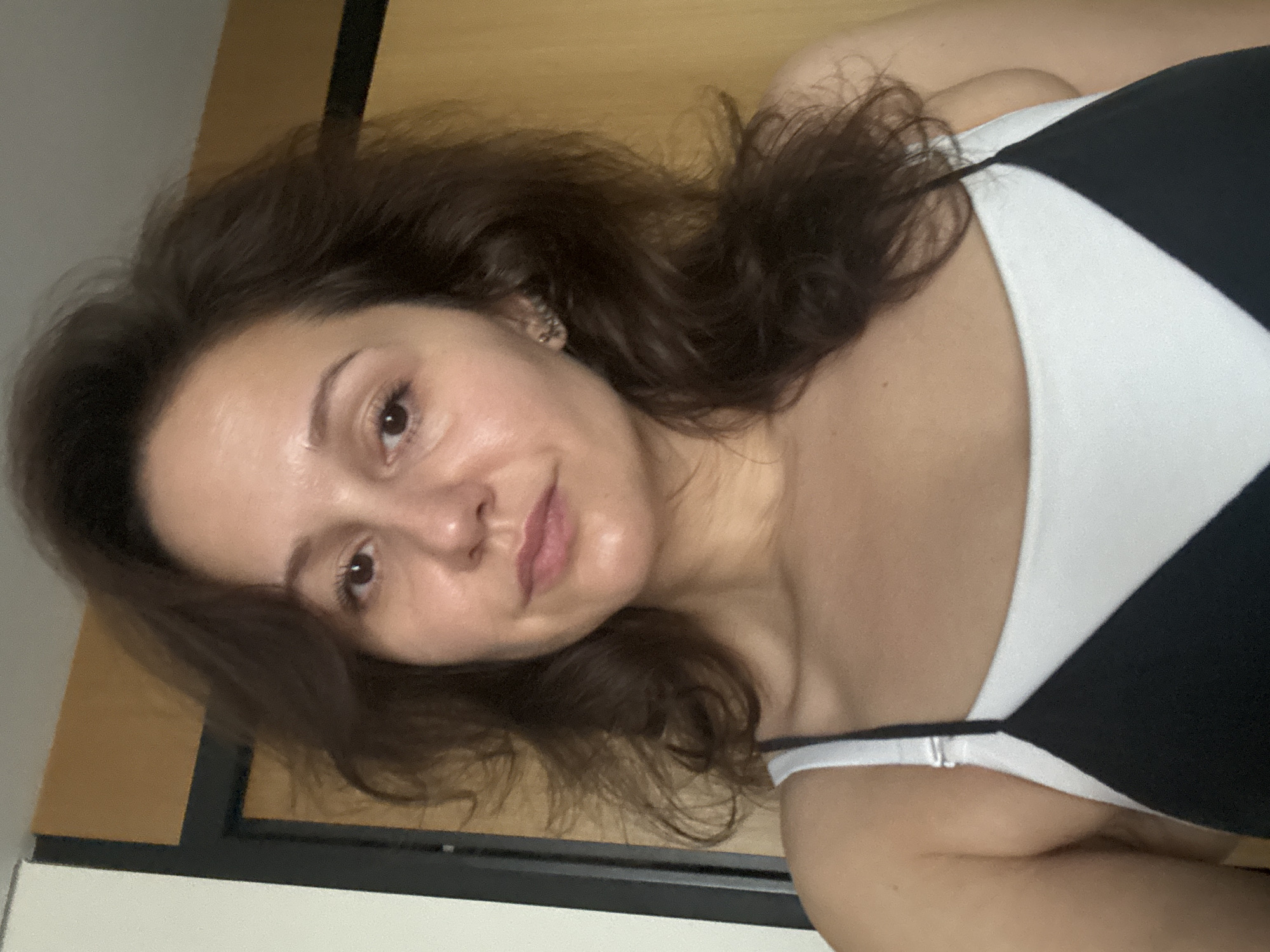 The image is a selfie of a woman with dark brown hair. She has light skin, brown eyes, and pink lips. She is wearing a black and white top. The background appears to be an interior setting with a door. The image does not provide any indication of the location being Dubai, United Arab Emirates.