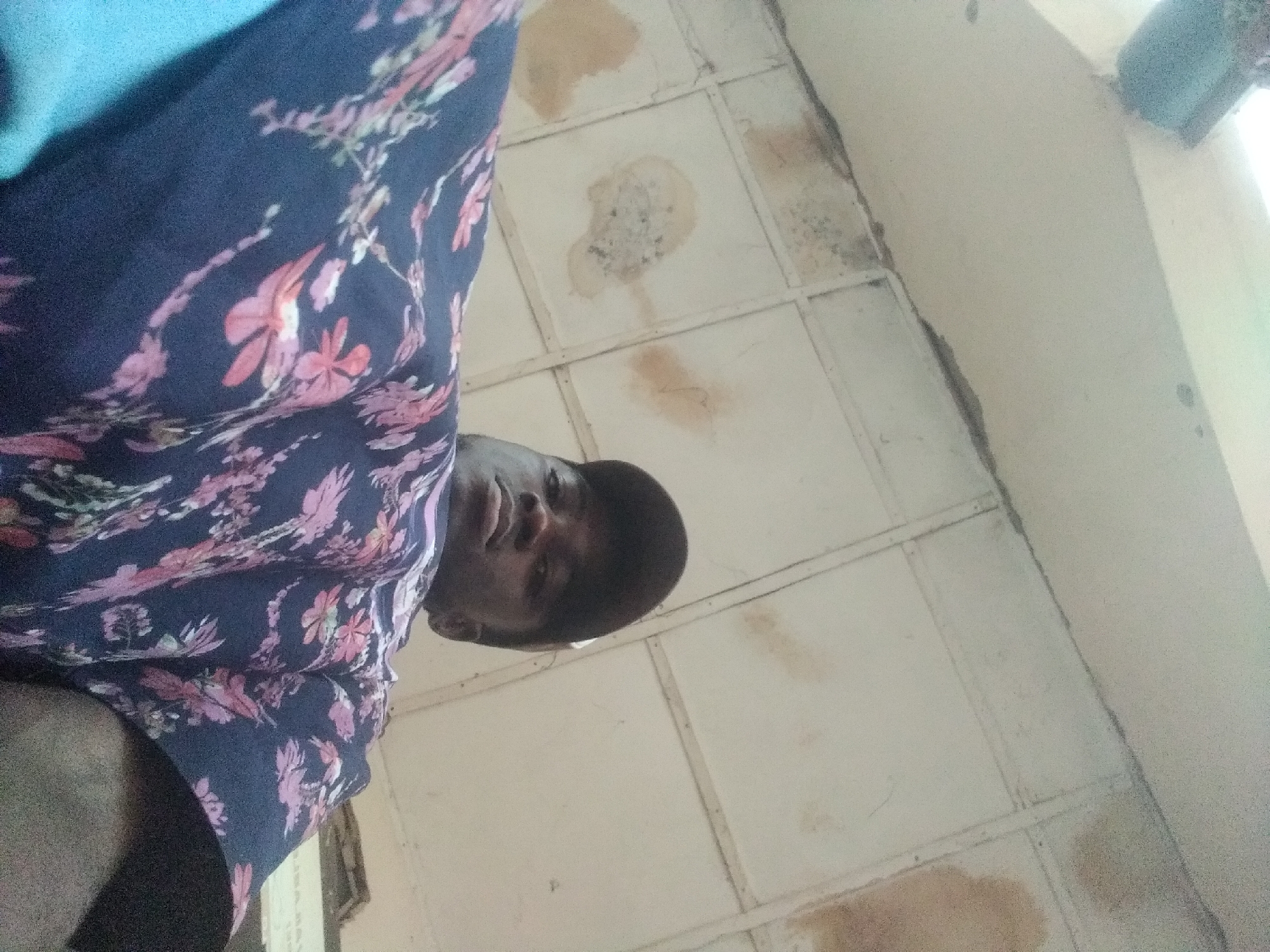 An overhead, low-angle selfie shows a young Black person wearing a dark baseball cap and a dark blue shirt with a repeating floral pattern in pink and white. The person is looking up towards the camera, and their face is partially obscured by the angle. The background appears to be a ceiling with a textured, off-white surface and visible water stains. The overall lighting is somewhat dim. The location is Jalingo, Nigeria.