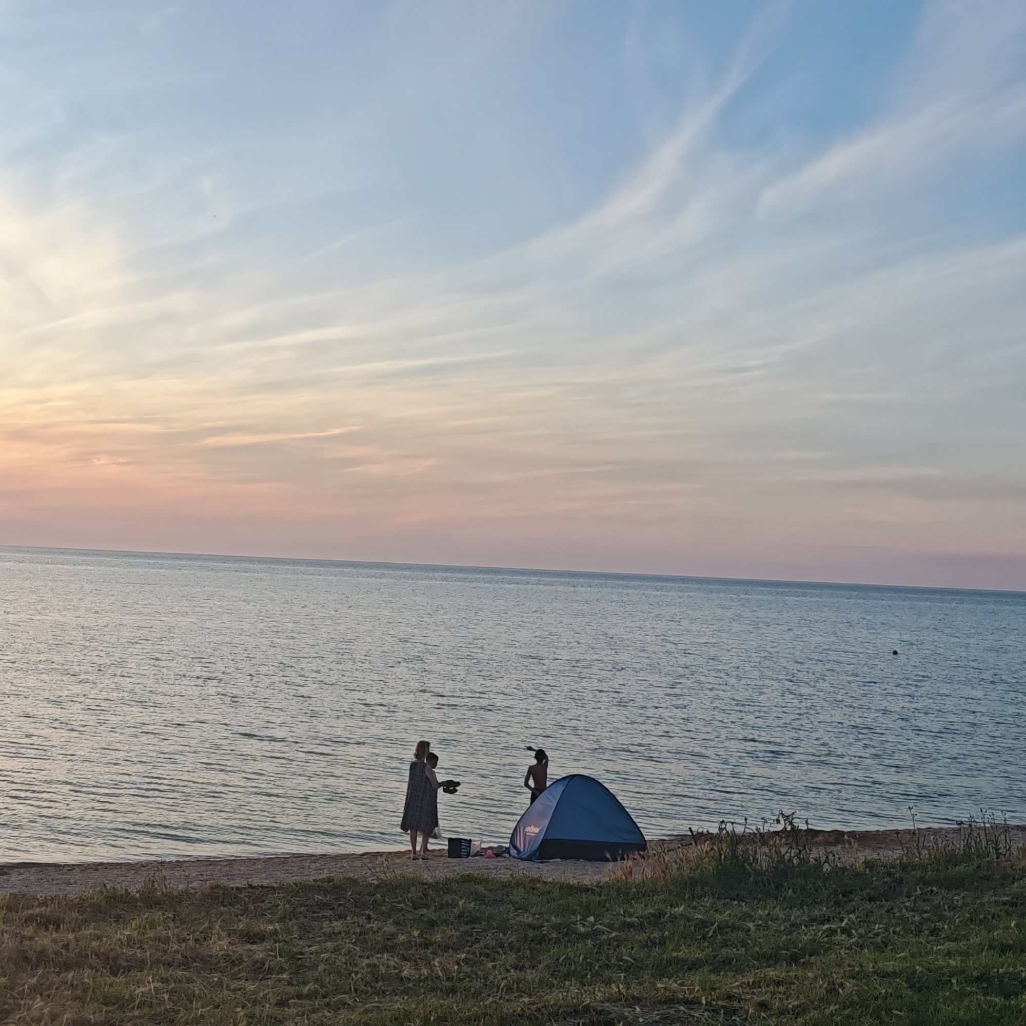 The image depicts a tranquil coastal scene at either sunrise or sunset, characterized by a soft gradient sky and calm water. The location is on a sandy beach in Unknown, Unknown.
The sky occupies the upper two-thirds of the frame, transitioning from a pale, light blue at the zenith to soft pink and peach hues near the horizon on the left. Wispy, elongated clouds are scattered across the sky, particularly in the bluer upper sections.
Below the sky lies a vast, calm body of water, likely a sea or large lake, stretching to the horizon. The water's surface is gently rippled, reflecting the muted light. A small, dark, circular object, possibly a buoy or a distant swimmer's head, is visible in the water in the mid-right section.
The foreground features a narrow strip of light-colored sand, constituting the beach, which meets a strip of wild, green and brown grass and low-lying vegetation in the immediate bottom foreground.
On the sandy beach, three individuals and a small tent are present. To the left, an adult female, dressed in a long, light-colored garment, stands holding a pair of sandals in her left hand. Beside her, a smaller individual, likely a child, stands wearing a light top and dark bottoms. Further to the right, a third person, appearing to be a young male, stands with his back partially to the viewer, shirtless and wearing dark bottoms, with his right arm raised and pointing towards the water or horizon.
Between the adult female and the pointing individual, a small, dark blue, dome-shaped tent is pitched directly on the sand. Adjacent to the tent, a dark bag or container and a pink object (possibly a towel or blanket) rest on the sand. The overall scene suggests a camping or leisurely activity on the beach during the golden hour.