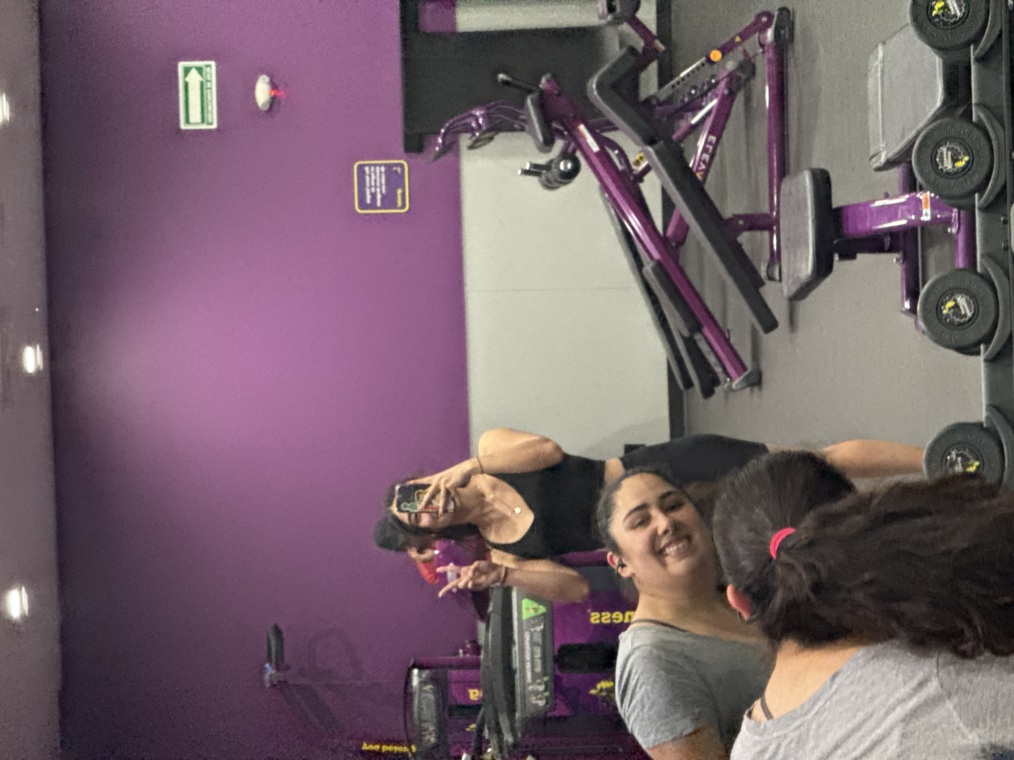 This image, originally rotated 90 degrees, captures an indoor gym scene in Puebla, Mexico. The setting is characterized by distinctive deep purple walls and purple and black exercise equipment, strongly indicating a Planet Fitness location.
Three women are visible. In the foreground, a woman with dark, curly hair tied back with a red hair tie is seen from behind, wearing a grey t-shirt. Standing behind her and slightly to the left, another woman with dark hair, wearing a grey t-shirt and earbuds, smiles brightly towards the camera. In the background, reflected in a large mirror, a third woman is taking a selfie with a smartphone, making a peace sign with her left hand. She wears a black tank top and a necklace, with sunglasses perched on her head.
The women appear to be casually socializing and taking photos during their gym visit. The atmosphere seems relaxed and friendly, conveyed by the smiles.
Visible details include various pieces of gym equipment, some with stacked weights. On the purple wall, a green rectangular sign with a white upward-pointing arrow is present, alongside a smaller yellow rectangular sign with indistinct symbols or text. A small white fixture with a red light is near the green sign. Text "2290" is visible on a piece of gym equipment in the reflection, and part of the phrase "pon pesos" can also be discerned, likely related to weight measurements or currency.