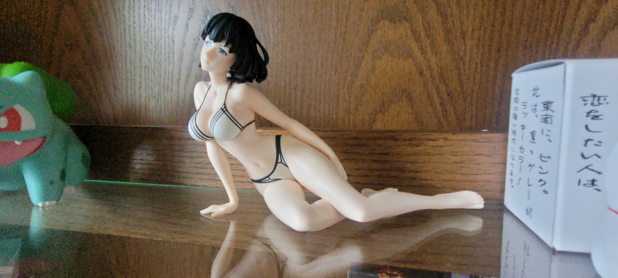 A figurine of a woman in a white bikini is displayed on a glass shelf in front of a wooden wall. To the left of the figurine is a green toy figure. A white box with Japanese writing is placed to the right of the figurine. The scene is likely indoors, possibly in a home or retail environment. There is no information to suggest the location is Heusenstamm, Germany.
