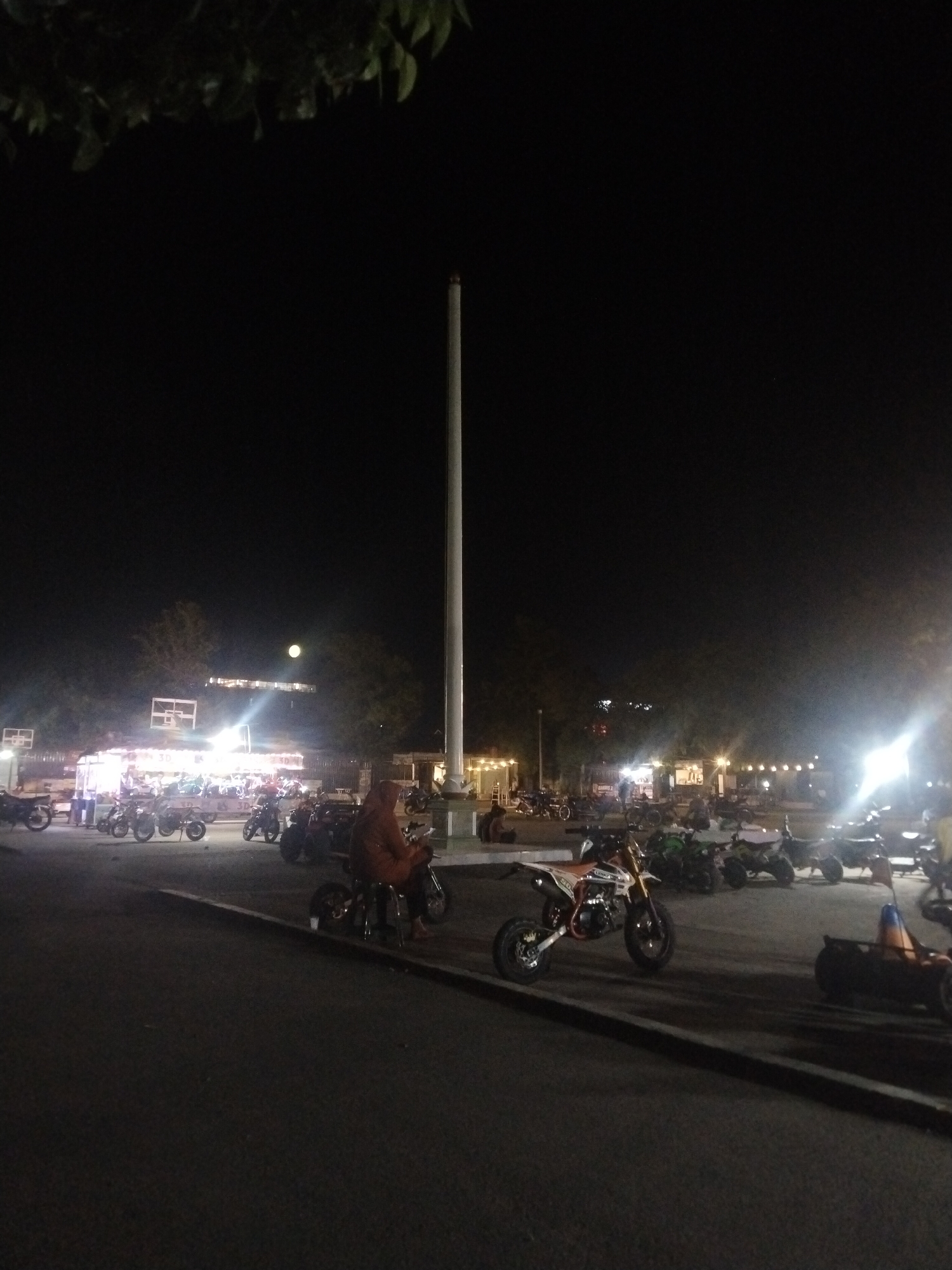 The image captures a bustling nighttime scene in what appears to be a public square or gathering area in Lhokseumawe, Indonesia. The sky is dark, illuminated by artificial lights and a prominent, full moon visible in the upper-left quadrant.
In the center-right of the frame, a very tall, slender white pole extends vertically into the dark sky, serving as a focal point. Below it, in the middle ground, are numerous brightly lit food stalls or vendor carts, creating a strong glow and attracting crowds. These lights illuminate various people gathering around, some standing, others seated.
The scene features many motorbikes, including several small, dirt bike-style or mini-bikes, suggesting a casual gathering or event involving motorcycle enthusiasts. In the immediate foreground, a person wearing an orange or brown hooded jacket is seated on a small motorbike, facing away from the camera. Other individuals are visible throughout the mid-ground, some interacting near the stalls, others sitting on their bikes.
The ground is paved, indicating an urban setting. Dark tree foliage is visible against the night sky at the top of the image and in the background. The overall atmosphere is lively and communal. On one of the brightly lit stalls, the text "3D" is vaguely discernible.