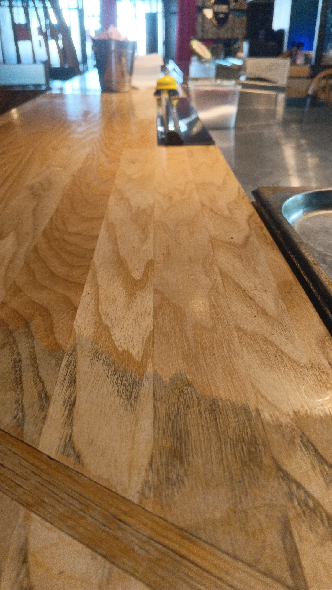 This is a close-up, eye-level, indoor shot of a wooden countertop with a section of a metal sink visible on the right. The wood grain is prominent, with variations in color and texture. In the background, out of focus, are glimpses of a bar or restaurant setting, including what appears to be a metal bucket with items inside, a yellow bell, and a stainless steel appliance. The lighting suggests ambient interior lighting. There are no people or discernible actions in the frame. The image does not contain any information about the city of Mytishchi, Russia.