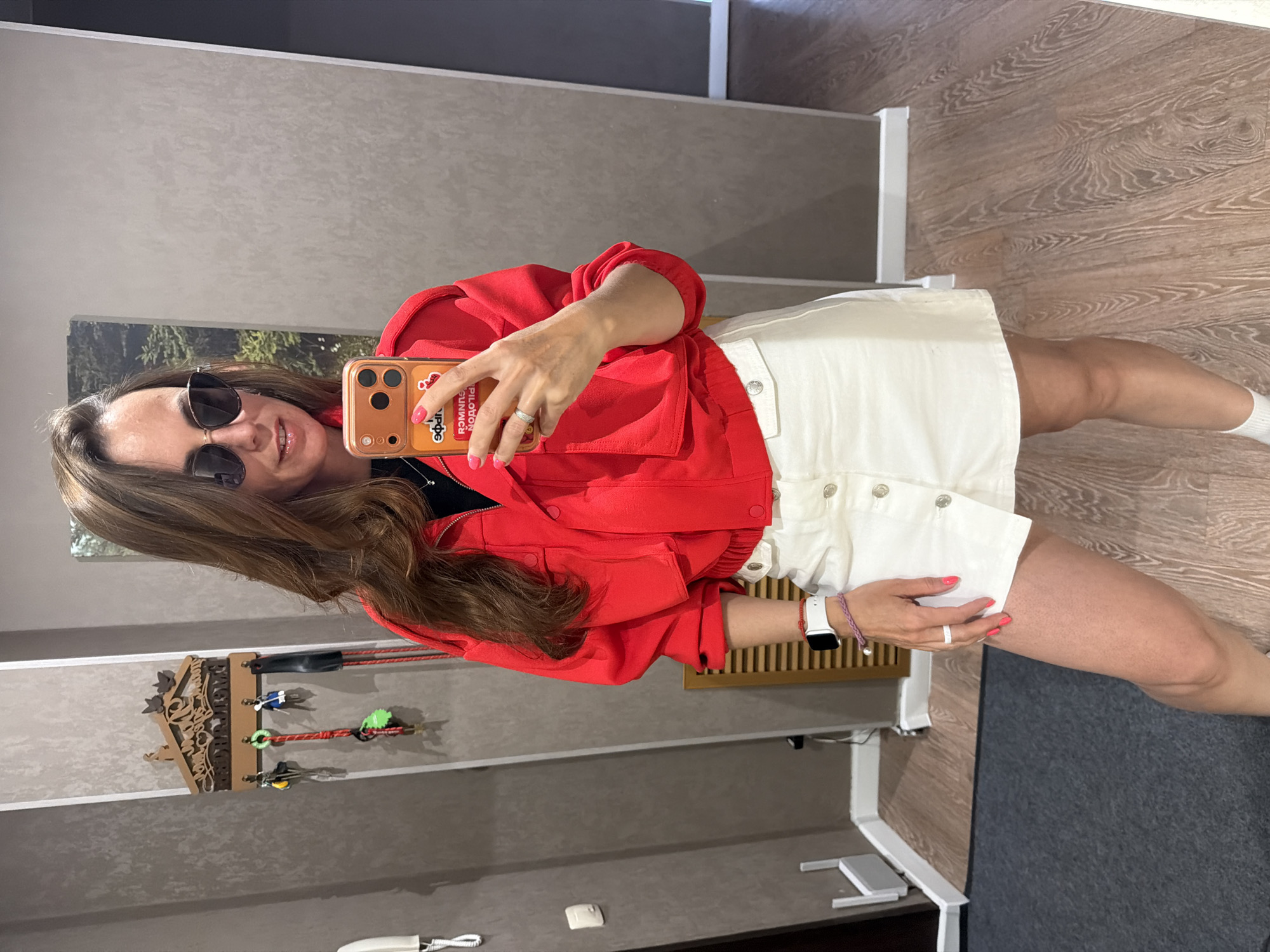 A woman is taking a selfie in what appears to be an indoor setting. She is wearing sunglasses, a red jacket, and a white skirt. She is holding a cell phone with a patterned case. The environment shows a mirror reflecting a key holder mounted on the wall, and a glimpse of an adjacent room with wooden flooring and a white door frame. No information indicates that this is Birobidzhan, Russia.