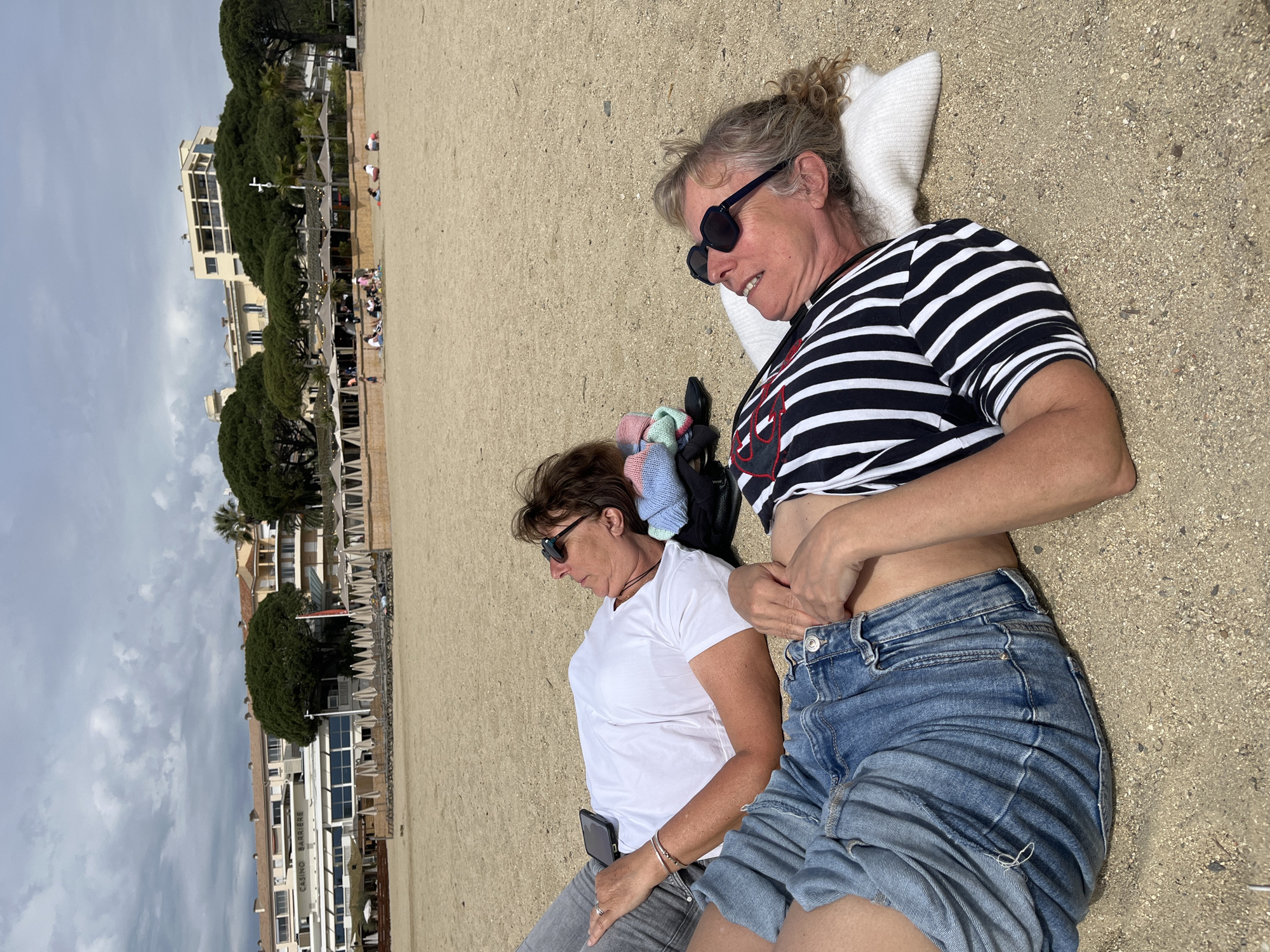 Two women are lying on a sandy beach on an overcast day. The woman closest to the camera is wearing a black and white striped shirt, jean shorts, and sunglasses. She is lying on a white pillow and has her hand on her abdomen near her unbuttoned shorts. The other woman is wearing a white t-shirt, gray shorts, and sunglasses. She is holding a cell phone. In the background are several buildings, including one with the words "Casino Barrière" printed on it, and some trees. The location is Sainte-Maxime, France.