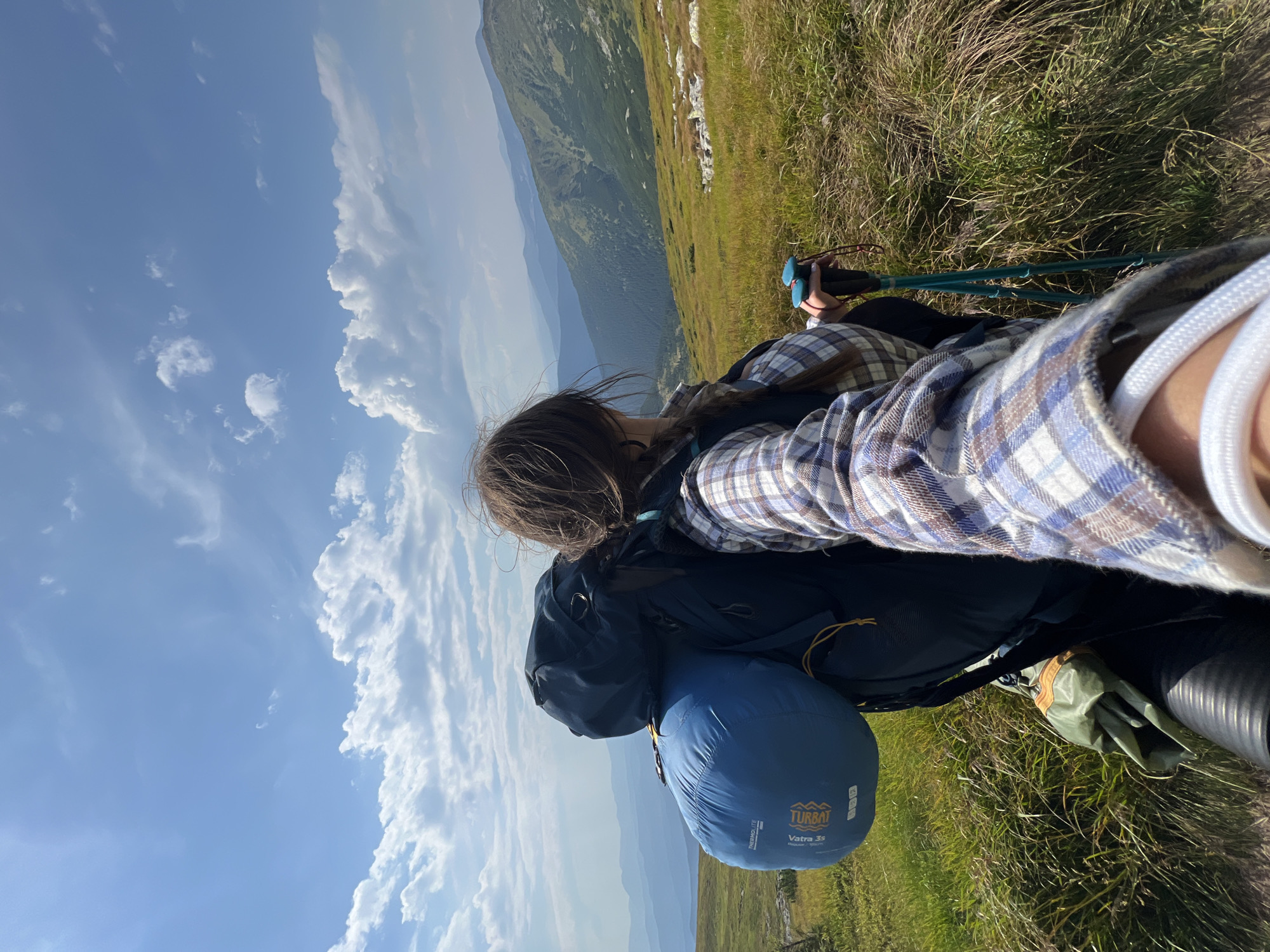 A person is taking a selfie while hiking in the mountains near Luhy, Ukraine. The person is wearing a plaid shirt and has a large backpack on. They are holding hiking poles in their hands. The background shows a scenic mountain landscape with green hills and a blue sky with white clouds. The sun is shining brightly.