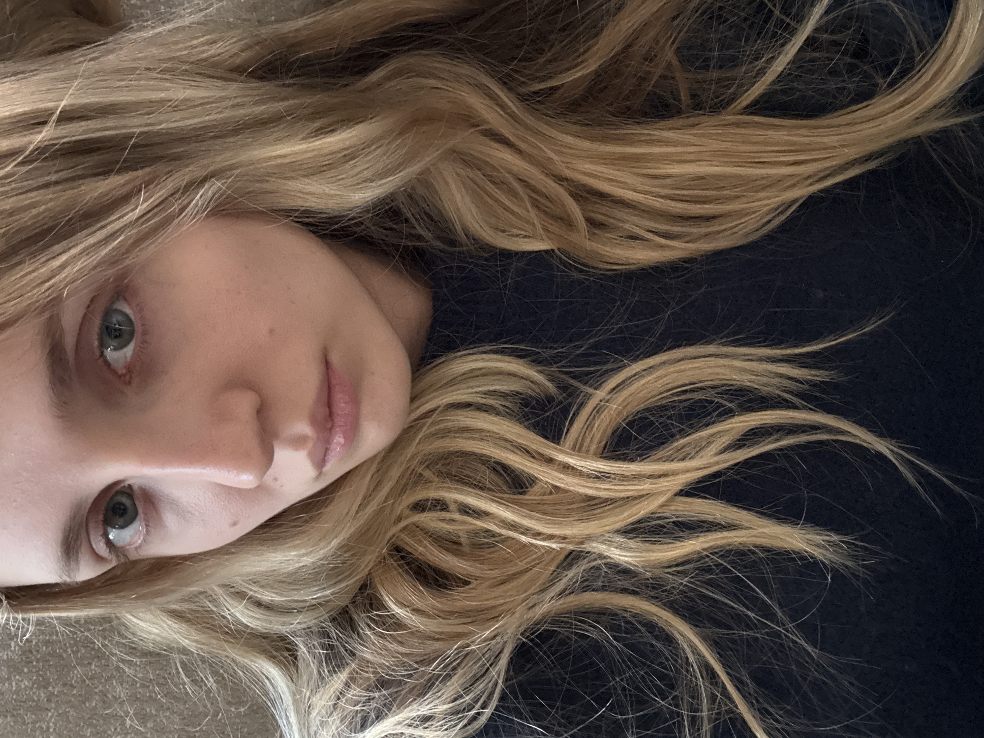 The image shows a woman lying down with her head turned to the side. Her hair is long, wavy, and blonde. She has fair skin, green eyes, and light pink lips. She is wearing a dark-colored shirt. The background is a plain dark color. There is no indication of the location being Mexico City, Mexico. There are no additional people, subjects, objects, actions, interactions, or environmental details beyond those described.