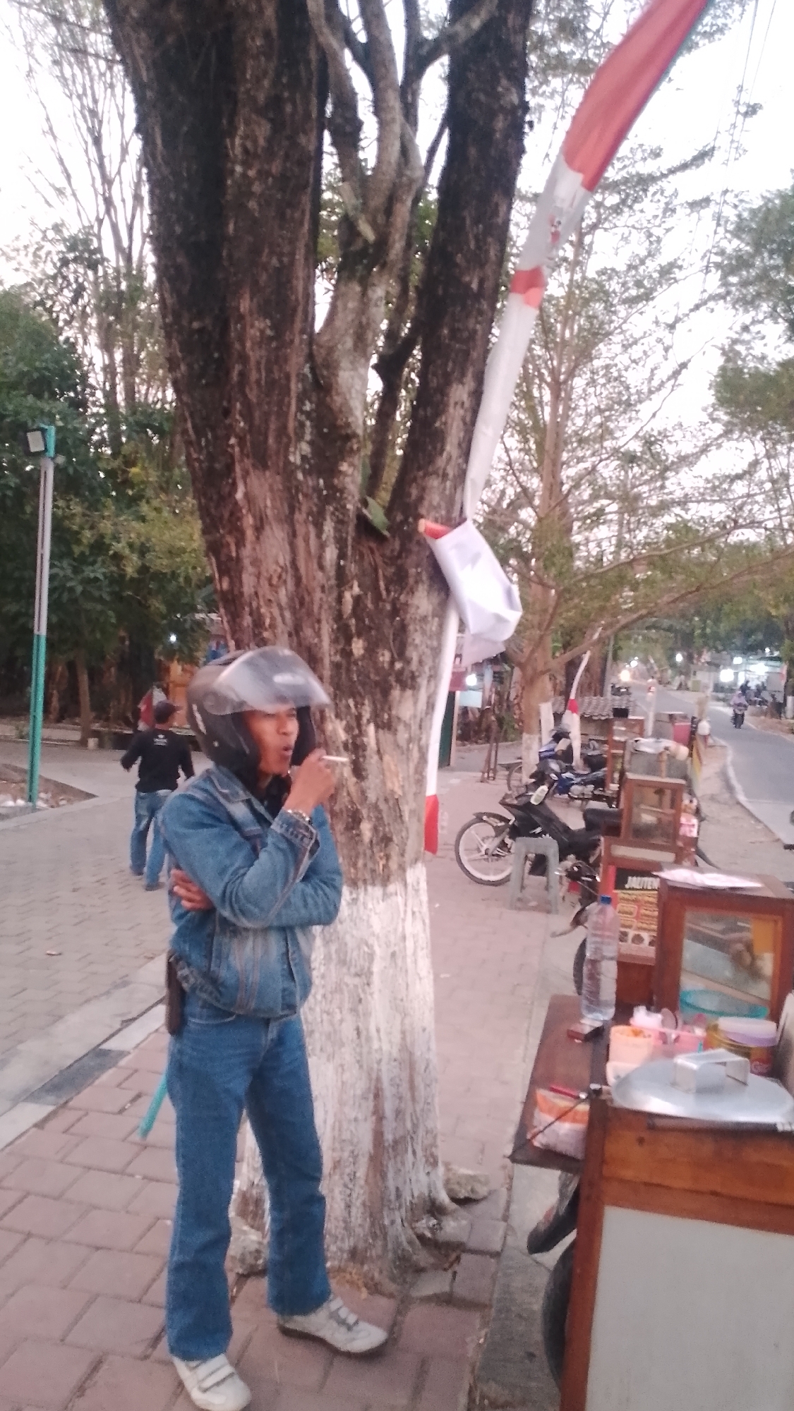 In the city of Karanglangu, Indonesia, a person wearing a denim jacket and jeans, with a helmet on, stands near a tree and is smoking. The person's right hand is raised to their mouth, holding a cigarette. Behind the person, there is a street with traffic and several parked motorcycles. A street food stall with wooden counters is visible to the right of the frame, displaying various items including bottles, containers, and a metal lid. The ground is paved with interlocking bricks. Trees and foliage line the background, with a pole and light fixture visible to the left. A red and white striped banner is attached to the tree.