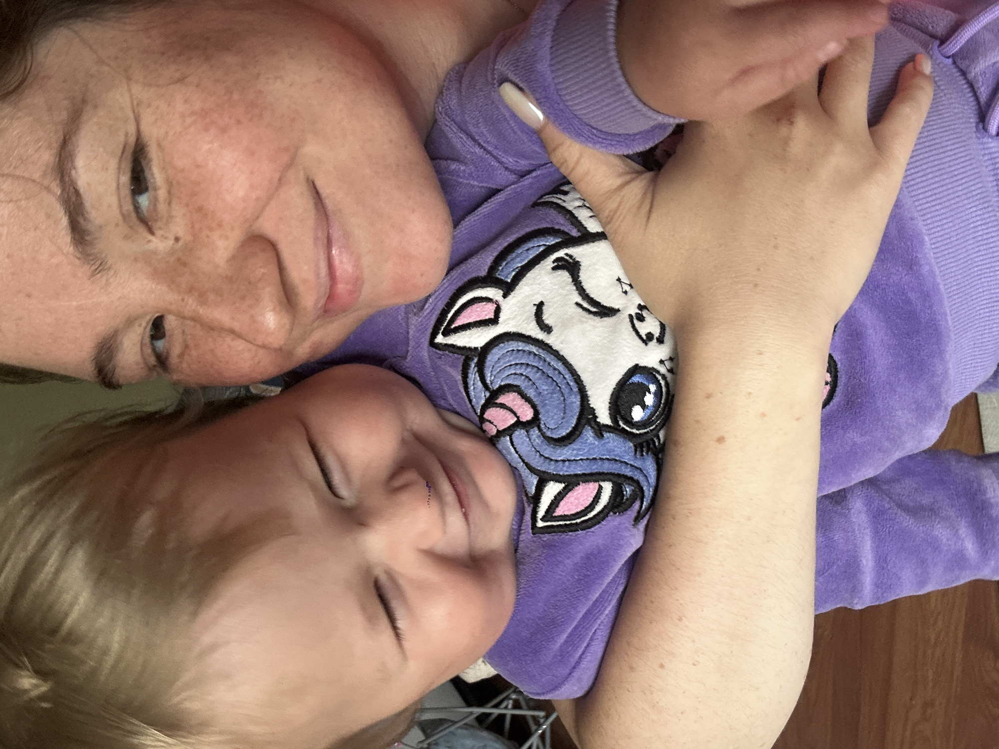 The image is a close-up, overhead shot of a woman and a baby, likely a mother and child, embracing. They are both wearing purple clothing. The baby is wearing a purple hooded sweatshirt with a cartoon unicorn design on the chest. The woman has freckles and is looking towards the camera with a slight smile. The baby is looking down and appears to be relaxed, with eyes closed and a content expression. Their heads are tilted, and the image is taken from a personal perspective, suggesting a candid moment. The setting appears to be indoors, with a wooden floor visible in the background. The lighting suggests daytime. There is no visible text in the image.