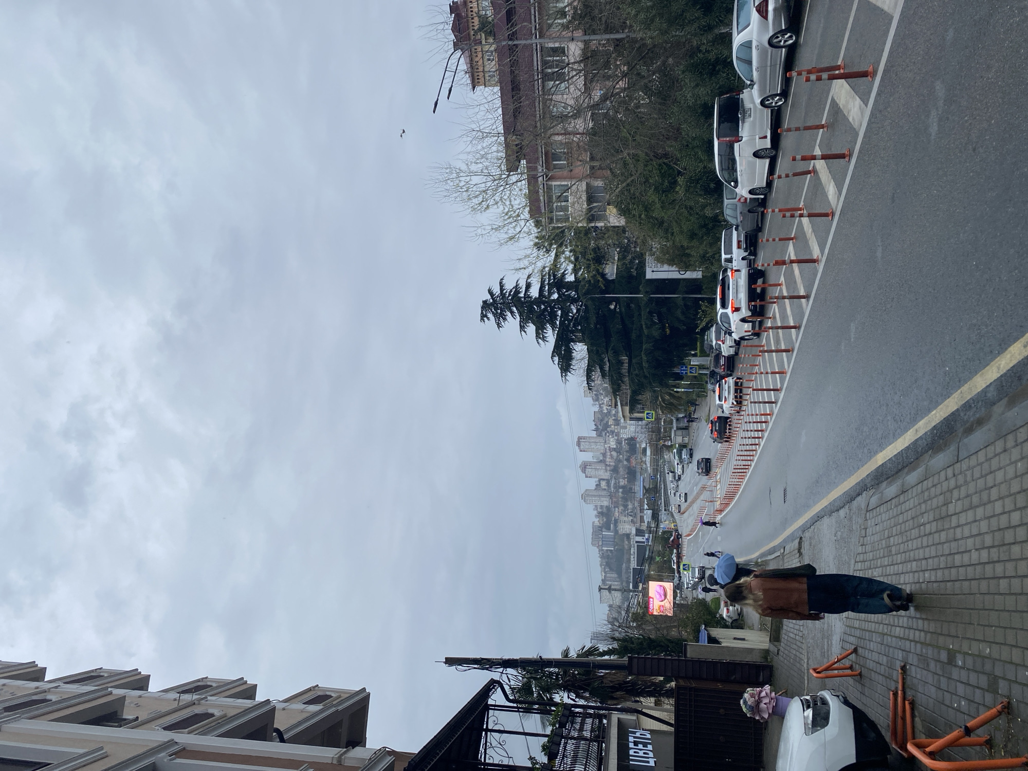 The image is taken from a slightly elevated position along a street in Sochi, Russia, on a cloudy day. The scene looks towards the city center, with a downward sloping road lined on both sides by parked cars and orange traffic cones. A woman with blonde hair, wearing a brown jacket and blue jeans, stands on the sidewalk with a blue umbrella. A parked white car with a bouquet of flowers on the hood is visible near a dark-colored building with a sign that reads "ЦВЕТЫ" (Flowers). Further down the road, the cityscape of Sochi is visible, with numerous high-rise buildings in the distance. The sky is overcast, and the overall atmosphere appears damp. Buildings of varying heights and styles line the street on both sides, with bare trees and foliage interspersed. Streetlights and power lines are visible above the street. A billboard is visible on the left. Traffic is moving on the road.