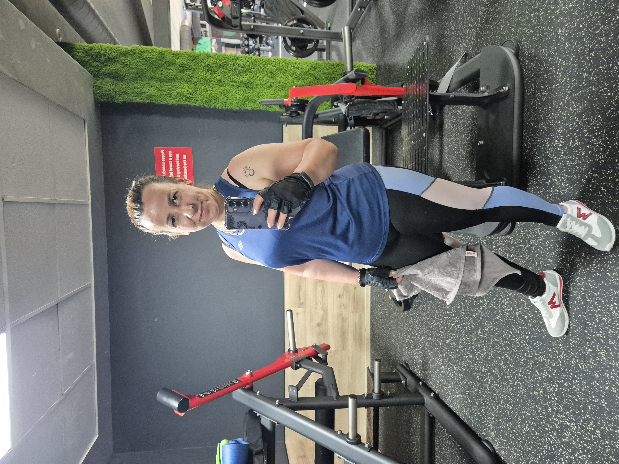 A woman is taking a mirror selfie in a gym. She is wearing a blue tank top, black and light blue leggings, black fingerless gloves, and white and gray athletic shoes. She is holding a gray towel in her left hand and a smartphone in her right hand. A tattoo of a sun and moon is visible on her left shoulder. The gym has dark gray walls, a black floor with white speckles, and various weight machines. A bright green, moss-covered pillar is located to the right of the woman. A red and gray weight machine is visible to her left. A sign with red lettering is partially visible on the wall behind her.
