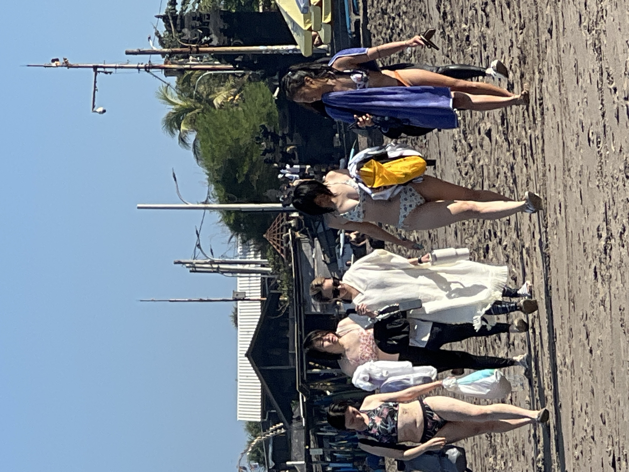Five women walk on a sandy beach near the city of North Kuta, Indonesia. The woman on the far left wears a floral bikini. The second woman from the left wears a pink bikini top and black pants, carrying a white bag. The third woman from the left wears a white cover-up and sunglasses. The fourth woman from the left wears a patterned bikini and carries a yellow bag. The woman on the far right wears a bikini top and orange bottoms, with a blue towel draped over her shoulder. In the background, there are various structures, including buildings, poles, and some vegetation. The sky is clear and blue.