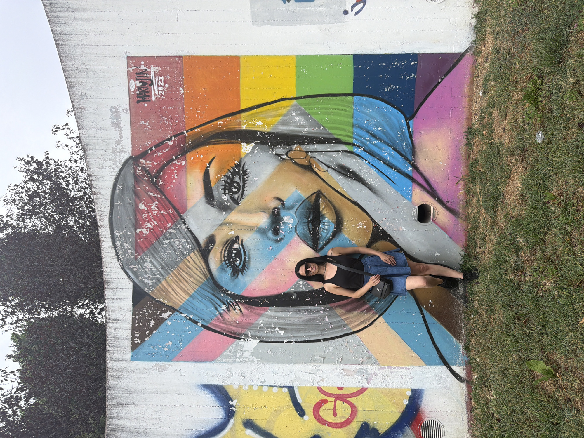 In Corsico, Italy, a woman stands before a large, colorful mural on a concrete wall. The mural depicts a stylized portrait of a woman's face, rendered with geometric shapes and a rainbow color scheme on the right side of her head. The woman in the foreground is wearing a black tank top, a denim skirt, black shoes, and sunglasses. She has a black crossbody bag slung over her shoulder. The wall itself is rough and textured, with sections of the paint visibly peeling. To the left of the main mural, another partially visible graffiti tag in yellow and red is present. The ground in front of the wall is covered in grass, with some patches of dry earth. Trees with dark foliage are visible above the wall. The sky is overcast.